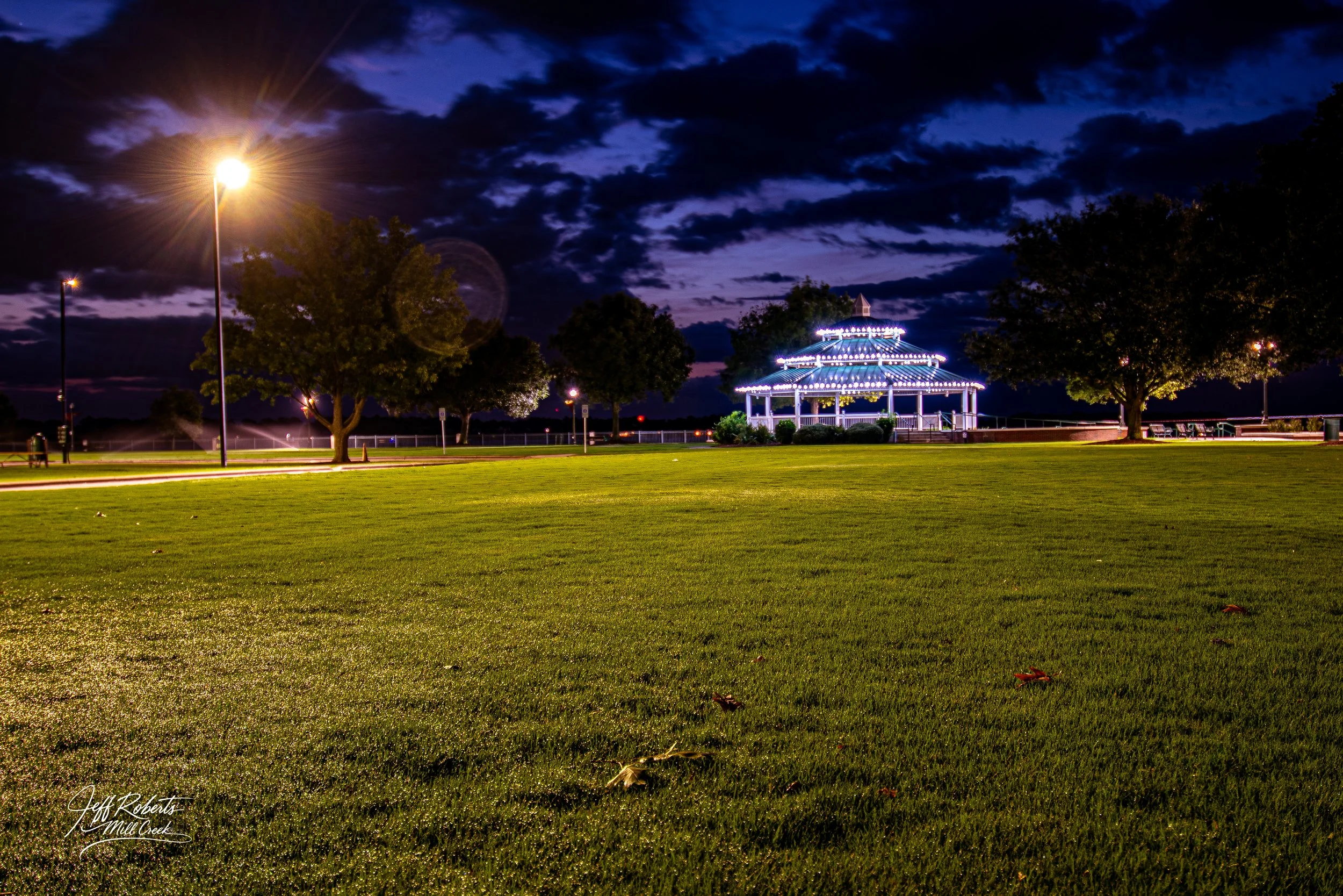 Nighttime park scene with illuminated gazebo, streetlights, trees, and a grassy field under a dark cloudy sky.