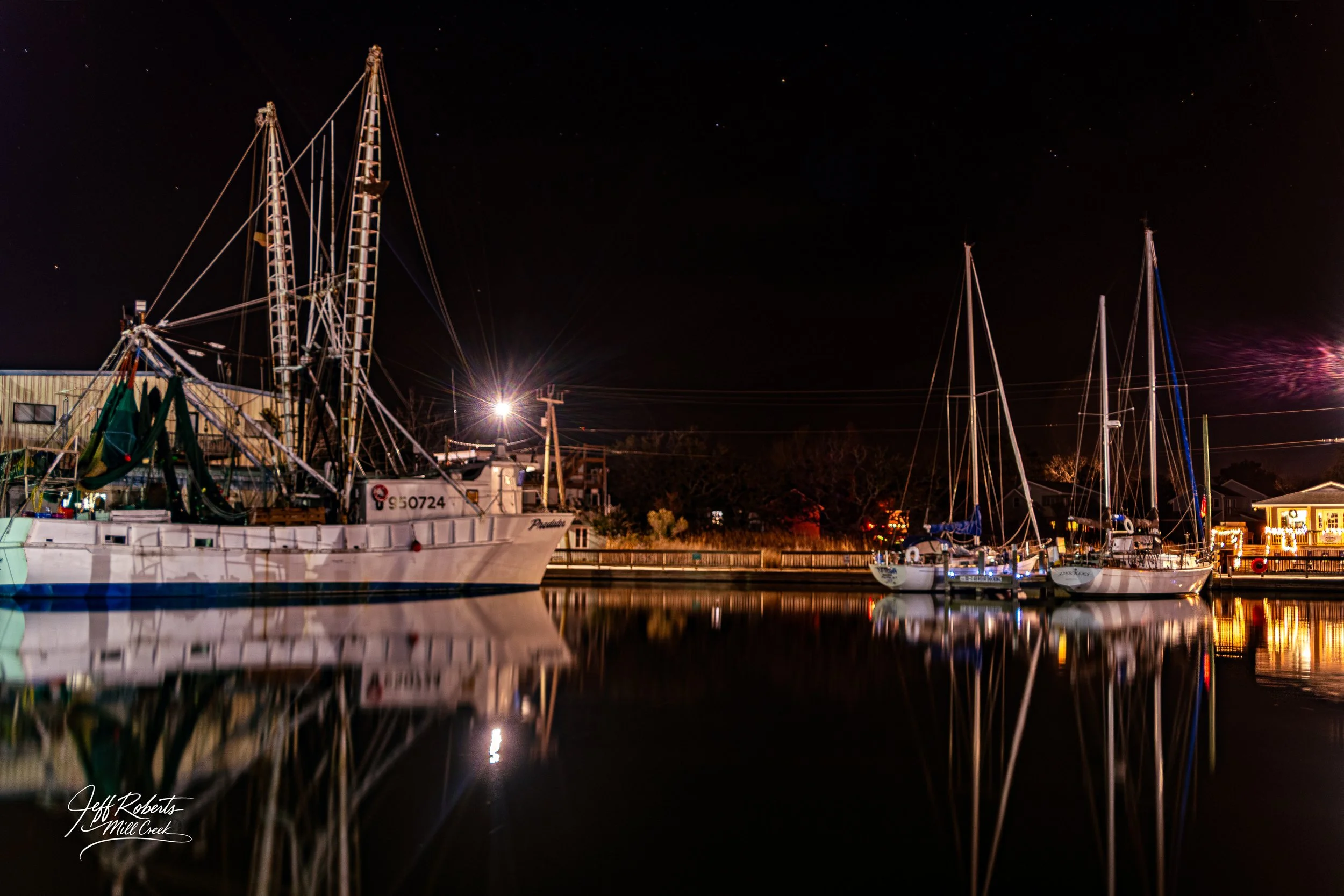 Nighttime view of sailboats docked at a marina with reflections on calm water, and buildings with lights in the background.