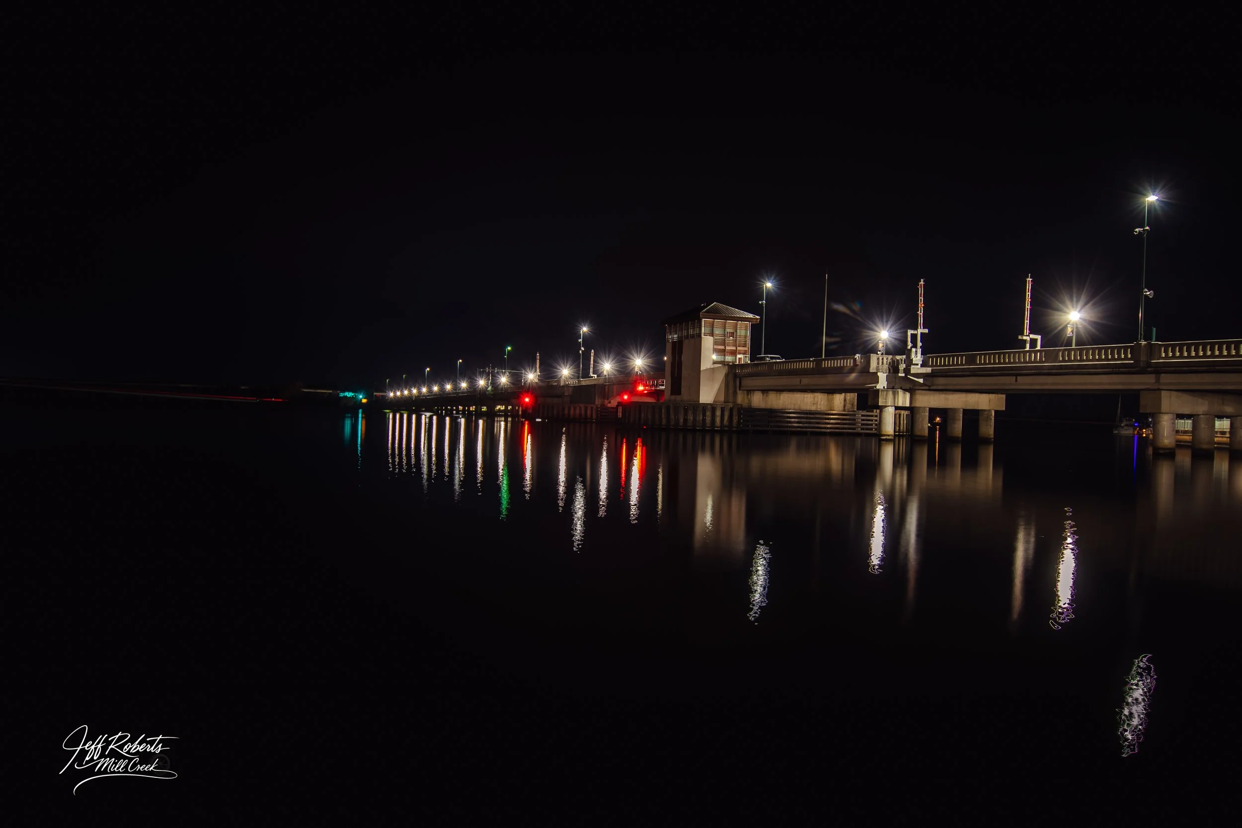 Night view of a lit bridge over water, with reflections of streetlights and traffic signals on the surface.