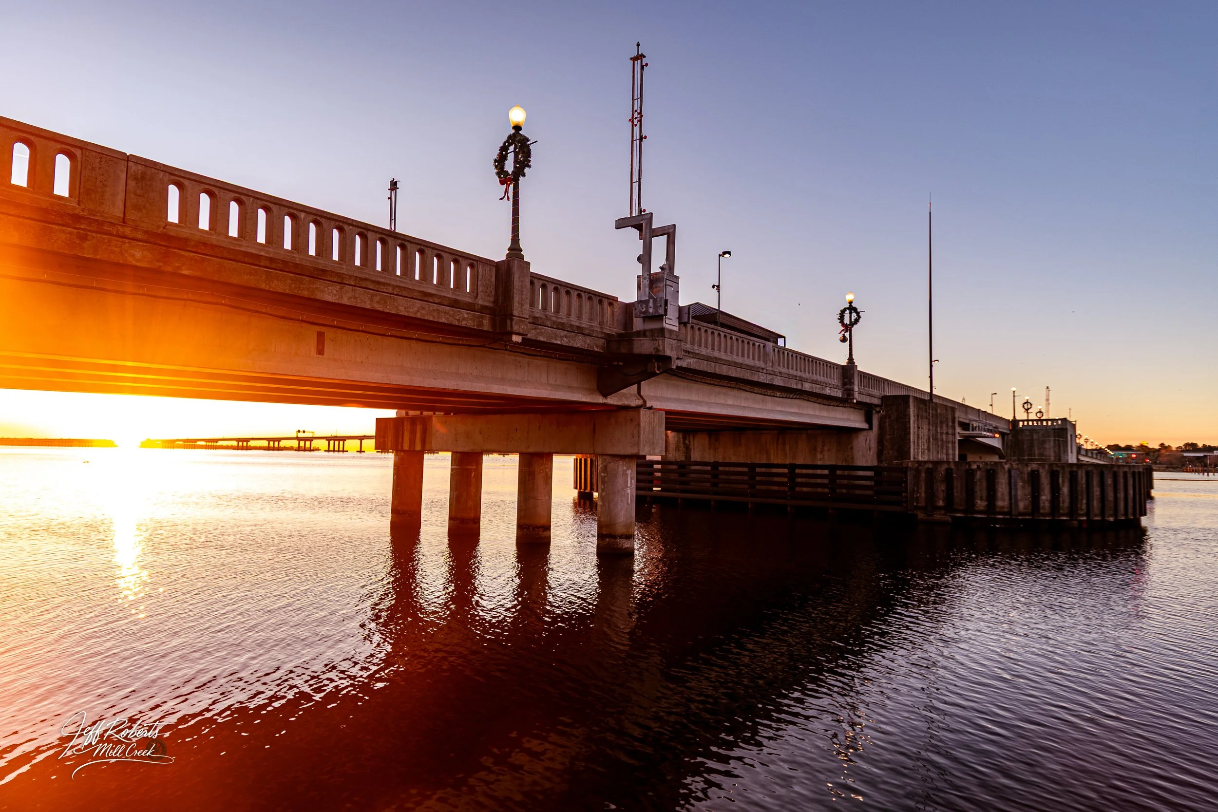 View of a bridge over water at sunset with holiday wreaths on lampposts