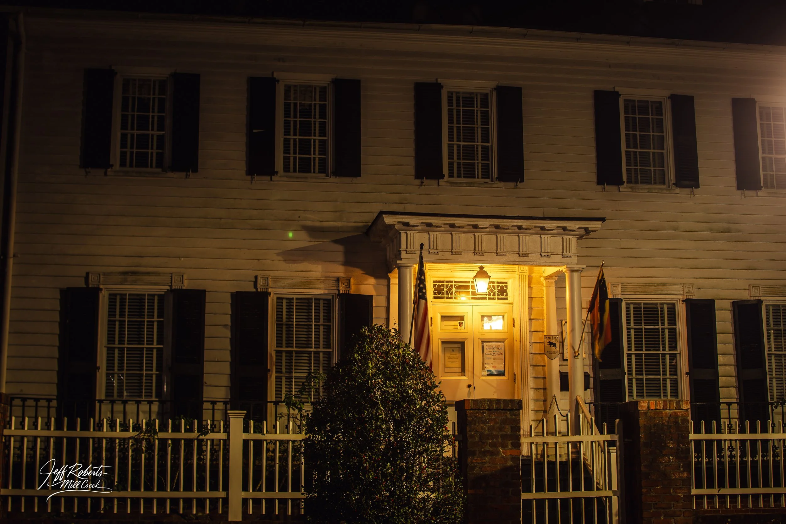 Nighttime view of a historic building with a lit entrance, black shutters on windows, white siding, and flags beside the door.