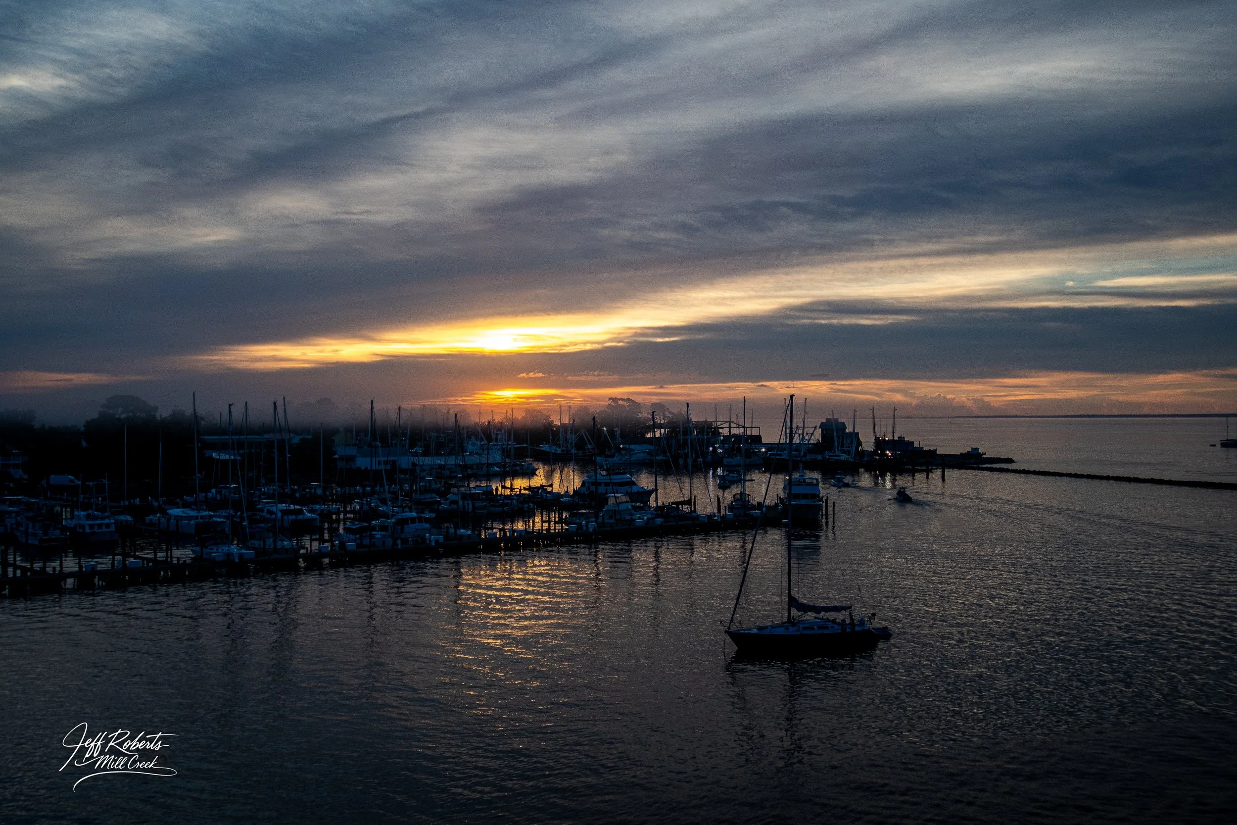 Sunset over a marina filled with sailboats and yachts, with calm water reflecting the sky and a boat sailing in the foreground.
