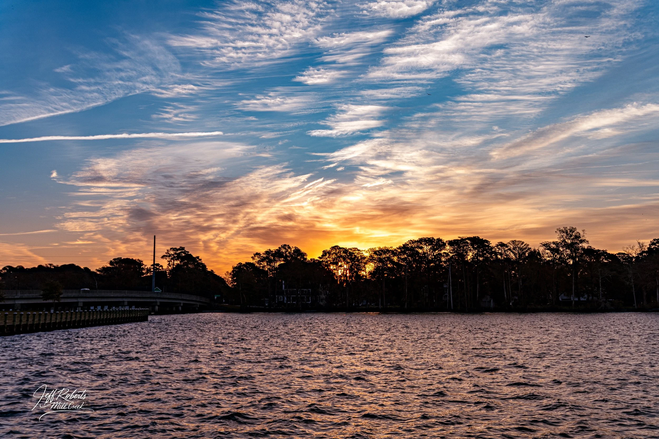 Sunset over a body of water with trees along the shoreline and a partly cloudy sky.