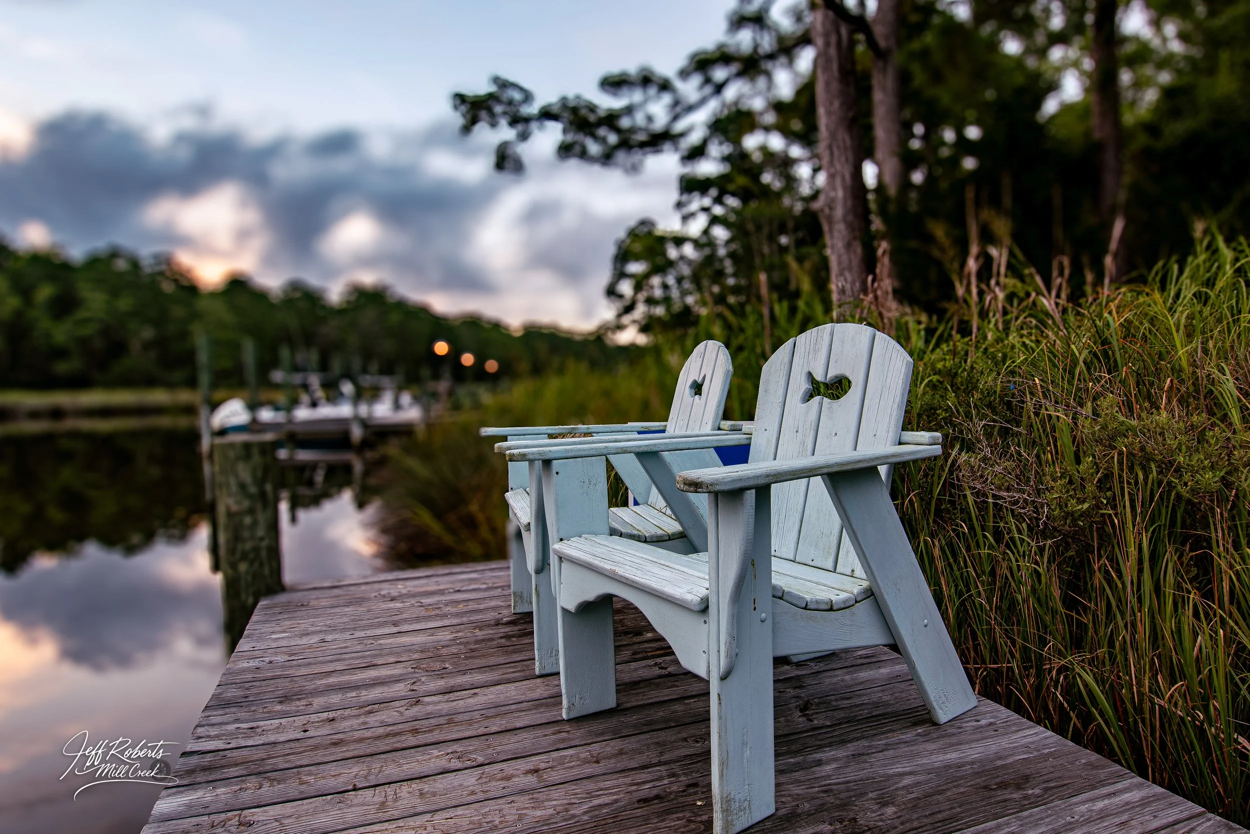 Two white Adirondack chairs on a wooden dock by a calm lake, with trees and a cloudy sky in the background.