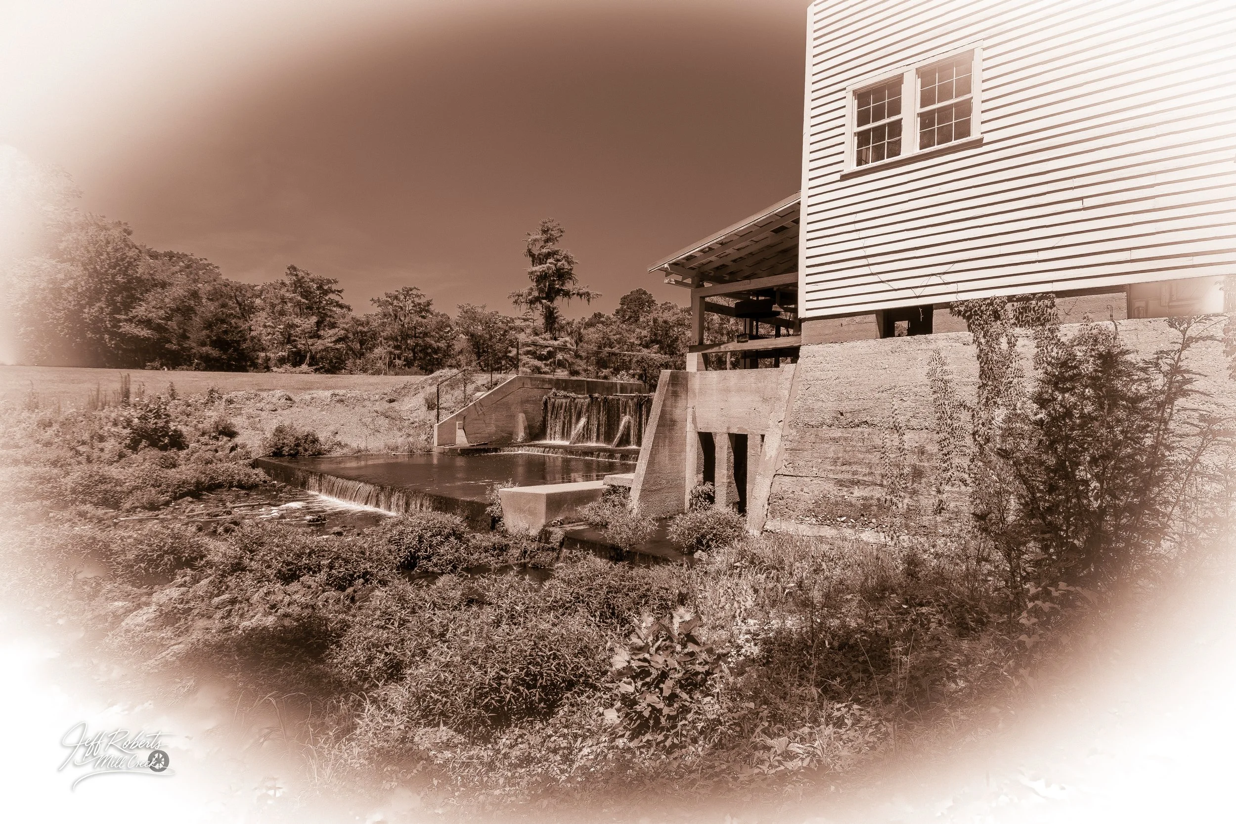 A sepia-toned image of a house on stilts beside a small waterfall and pond with trees in the background.