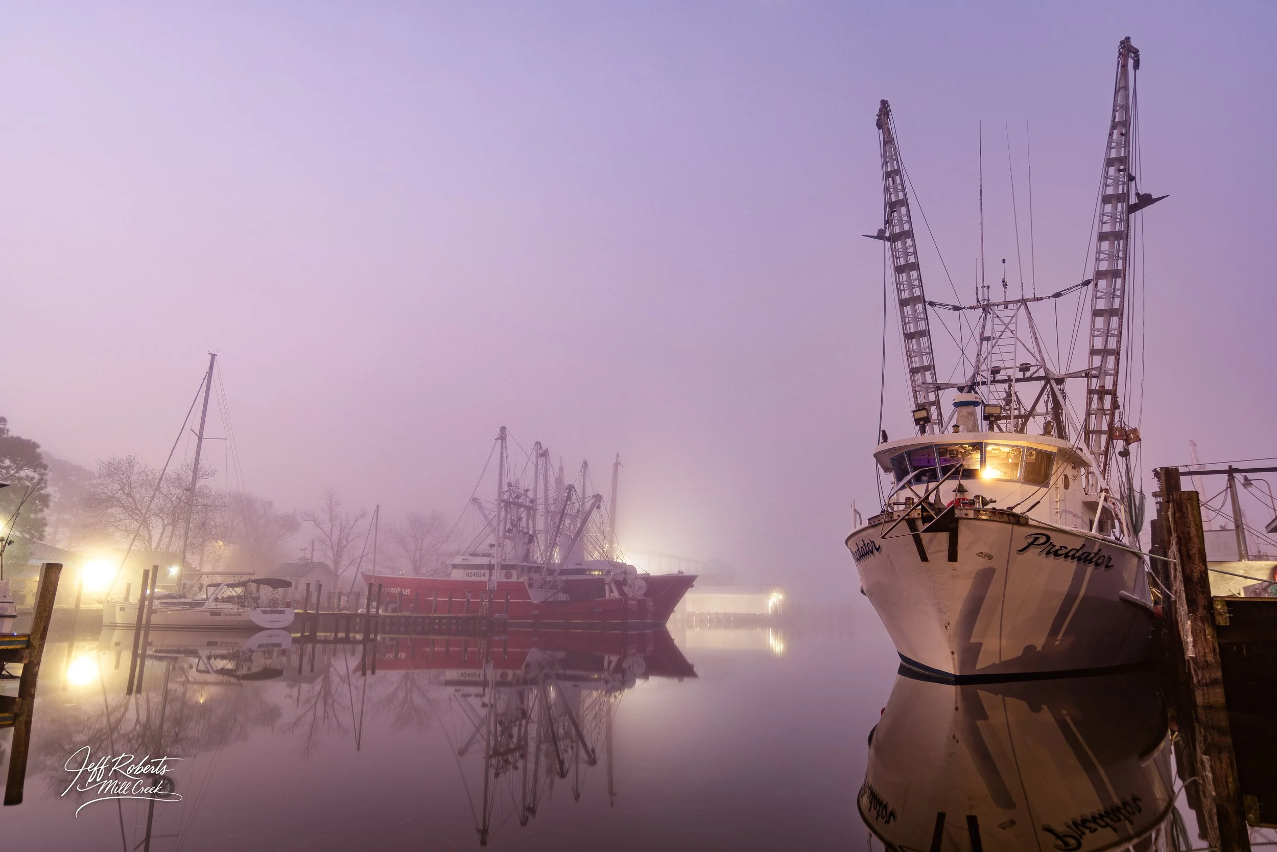Calm foggy morning at a marina with boats docked, their reflections visible in the still water, soft lights illuminating the scene.