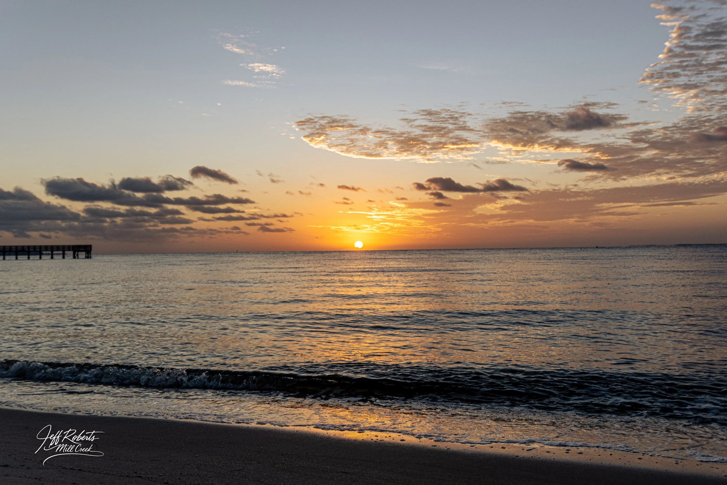 Sunset over the ocean with clouds in the sky and a shoreline with gentle waves in the foreground.