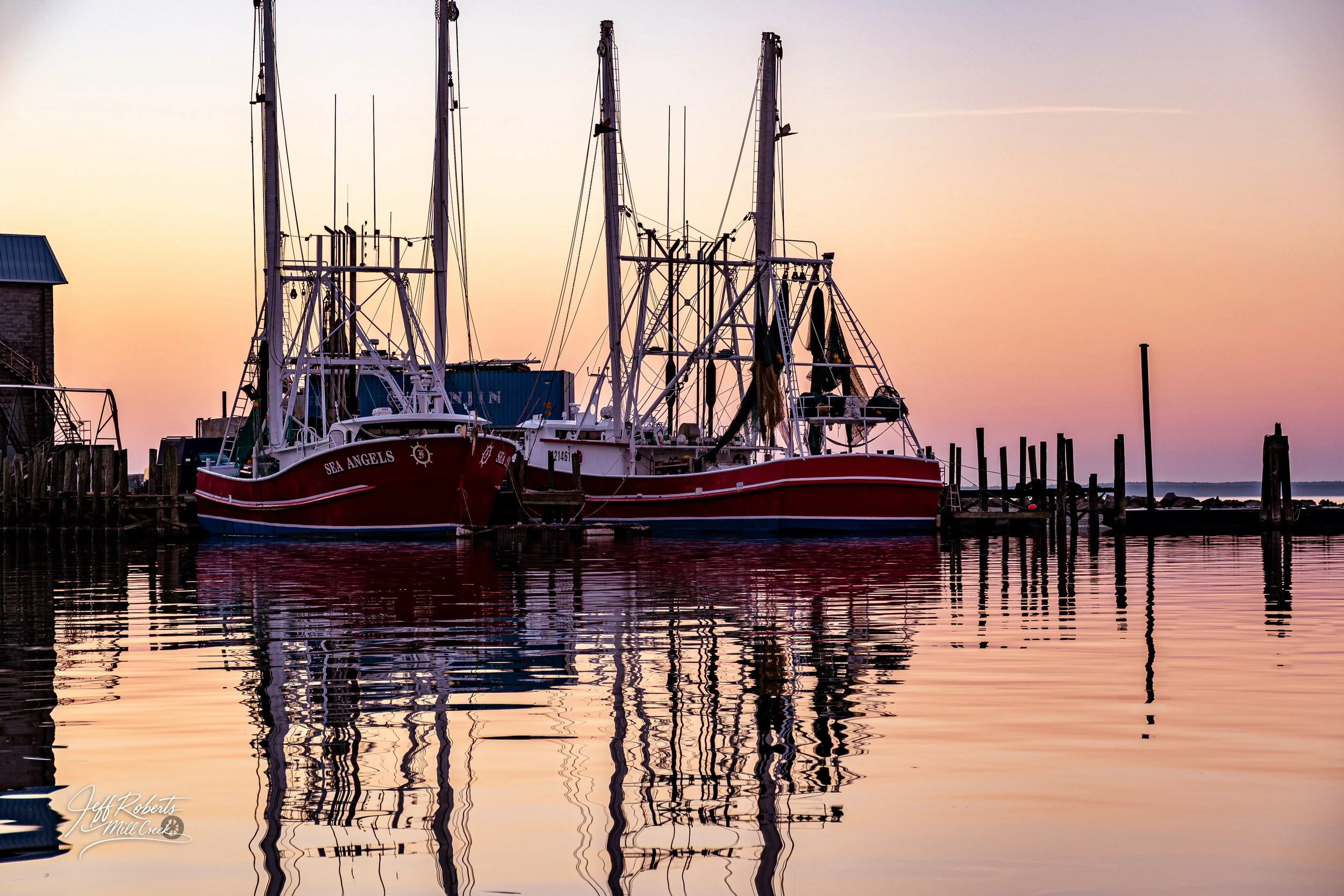 Two red and white sailboats docked at a pier during sunset, with reflections on calm water.