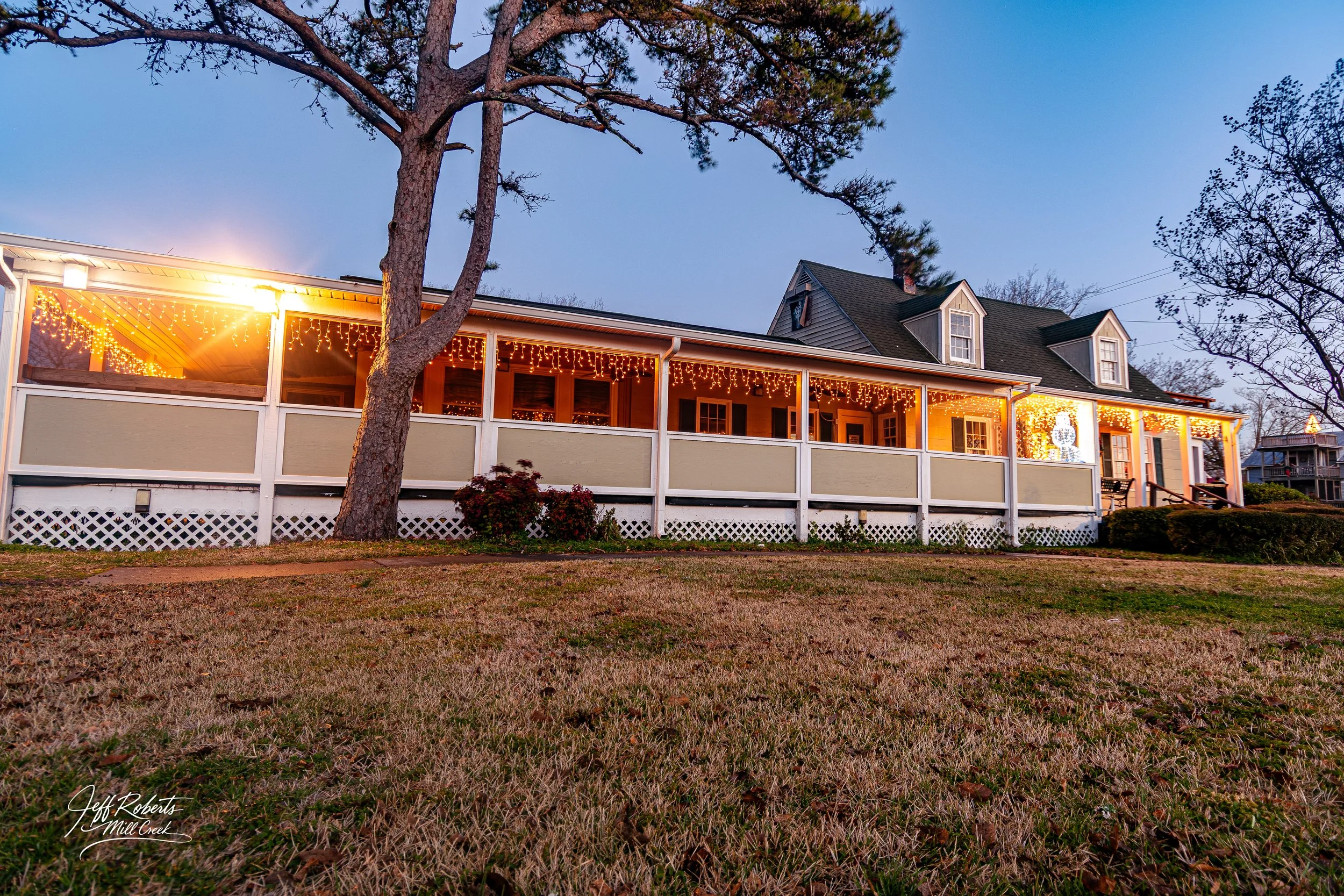 A house with a covered porch decorated with string lights, situated on a grassy area with trees and a clear evening sky.