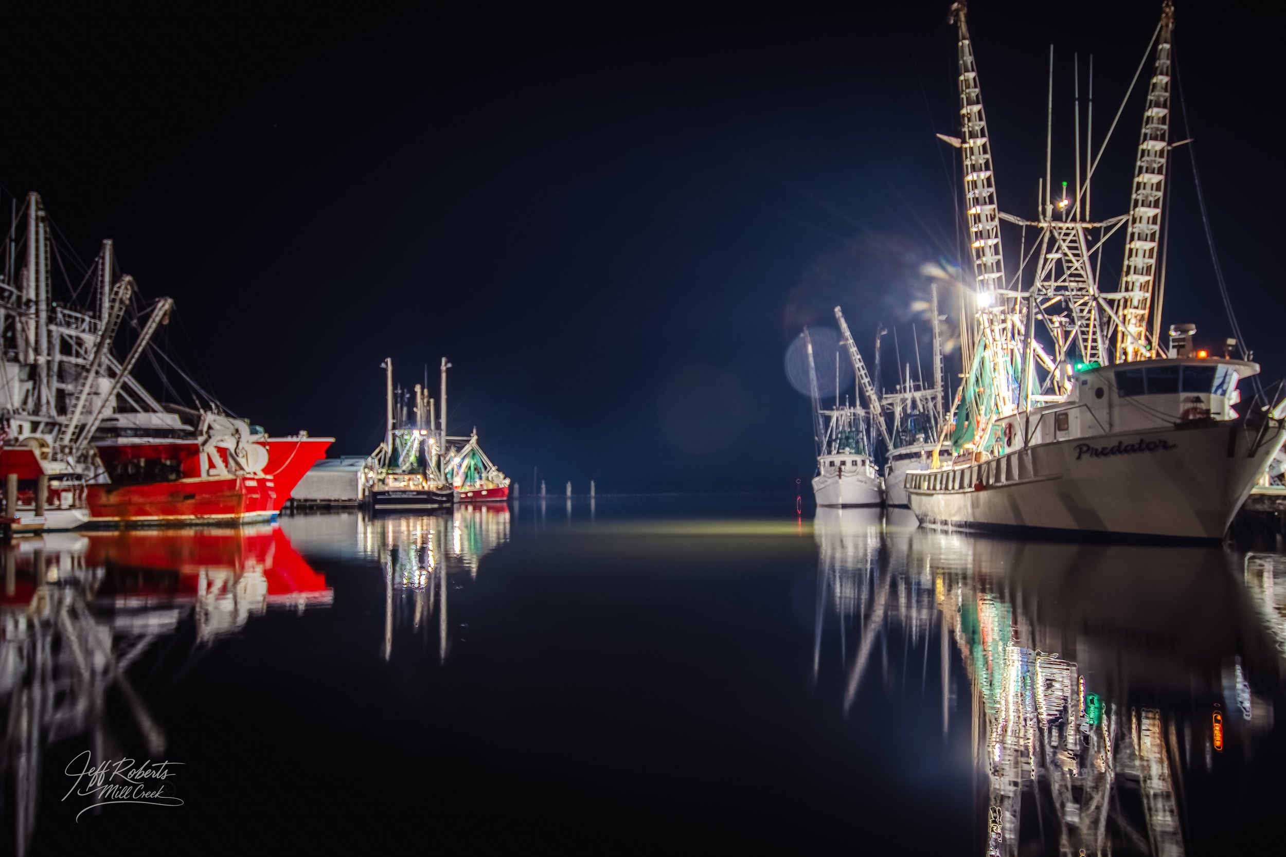 Night view of docked boats and ships at a harbor, with reflections on calm water and bright lights illuminating the vessels