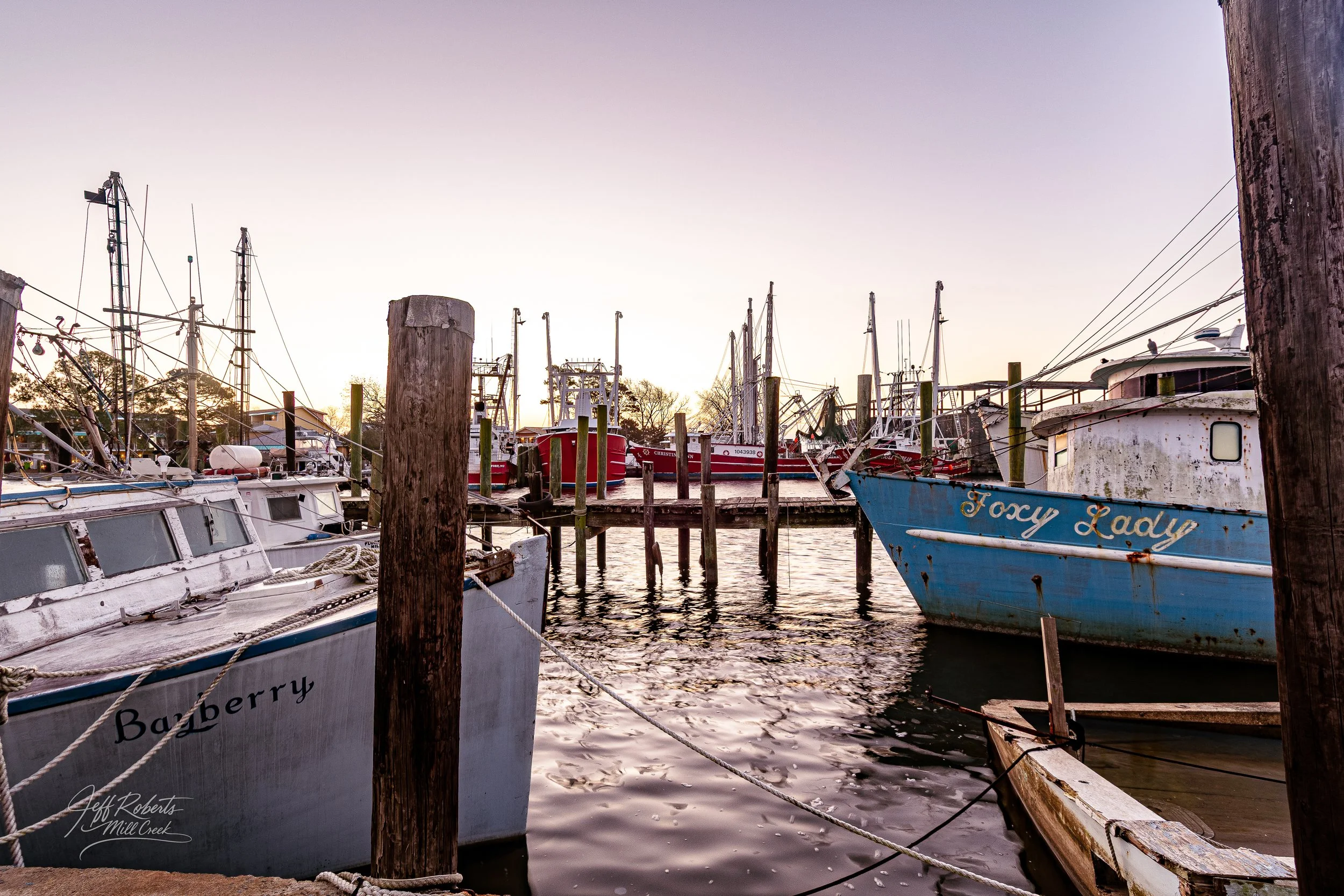 Several boats docked at a marina during sunset, with wooden posts and calm water.