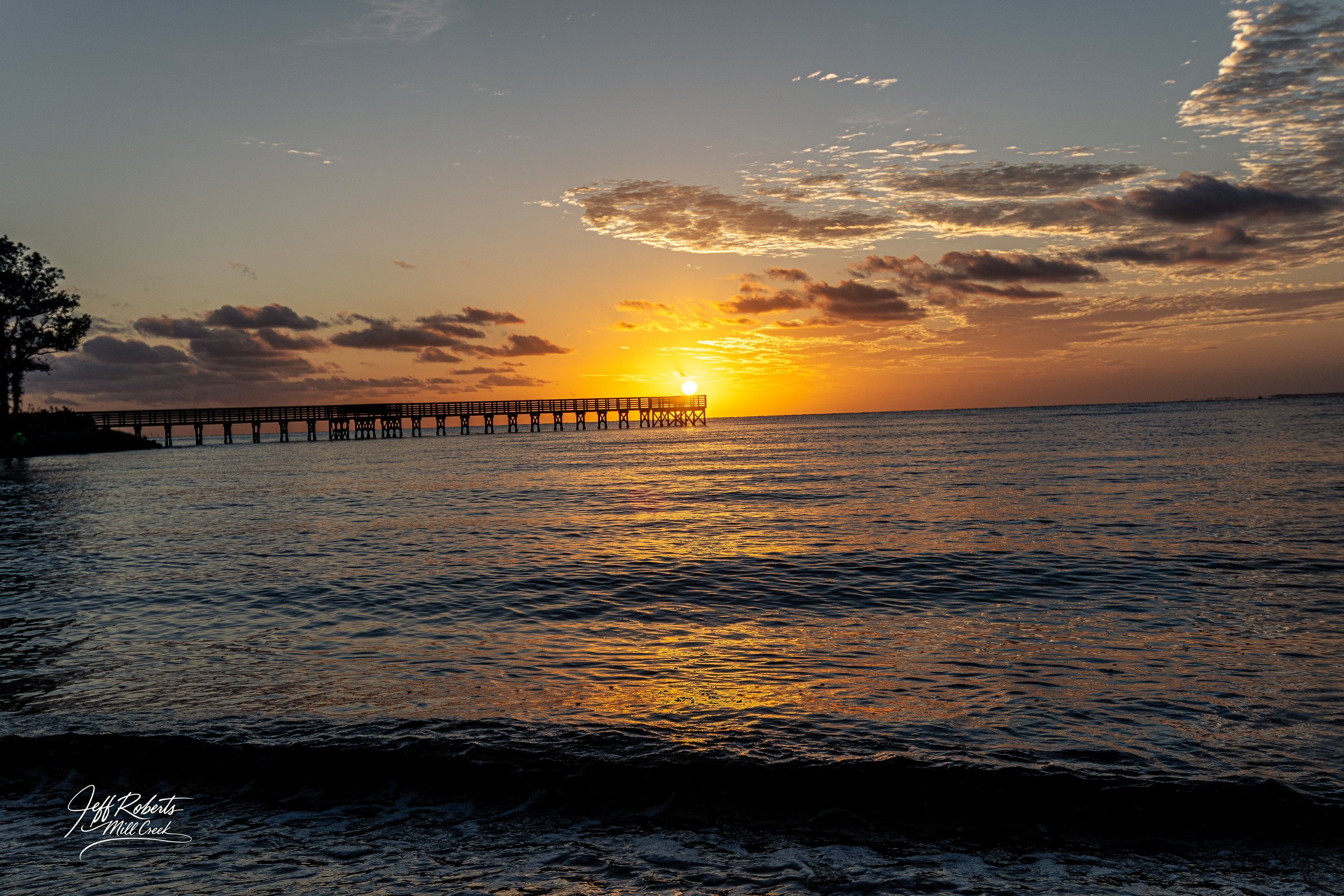 Sunset over the ocean with a pier extending out into the water, some clouds in the sky, and reflections on the water's surface.
