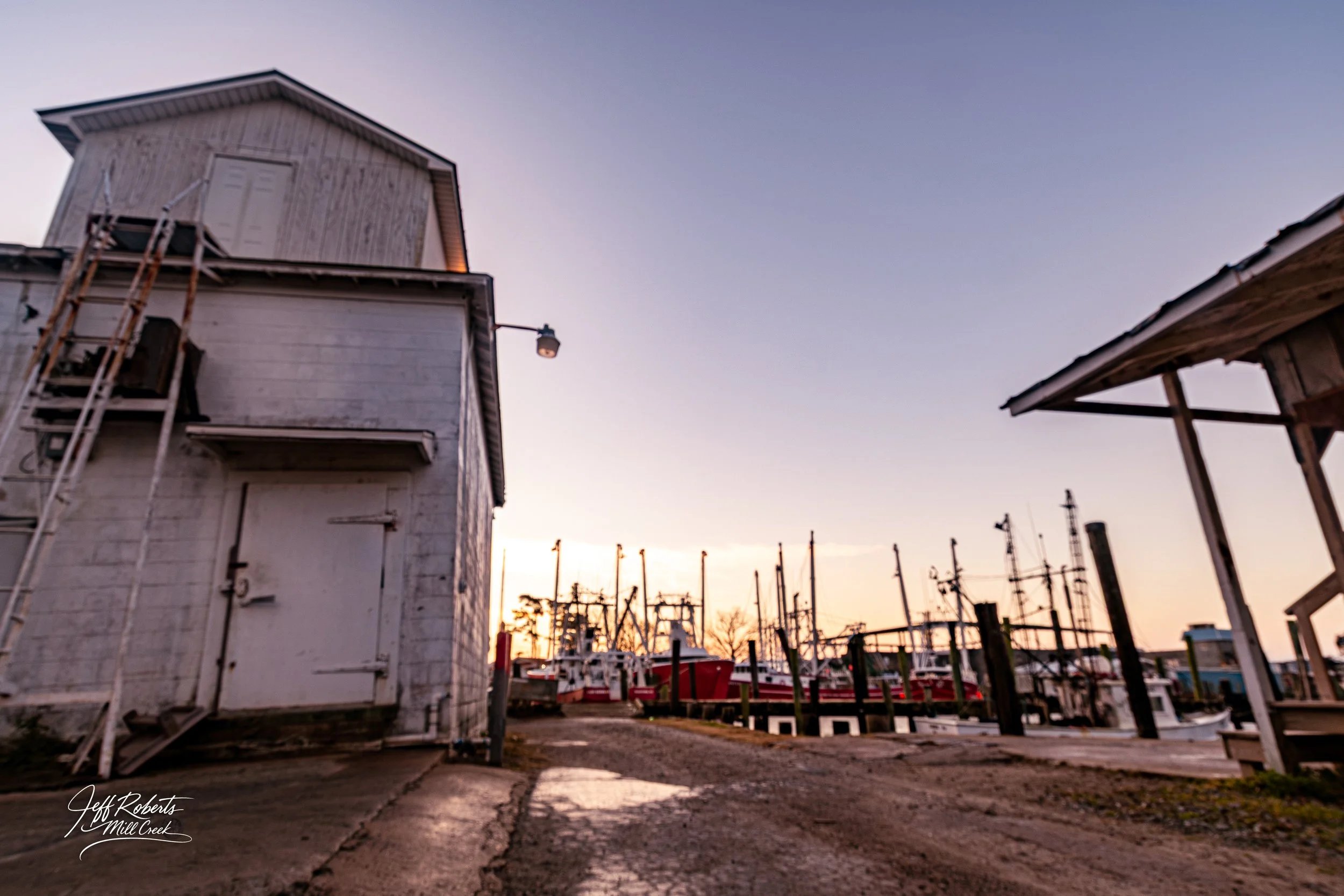 Fishing boats docked at a marina during sunset, with a small white building in the foreground.