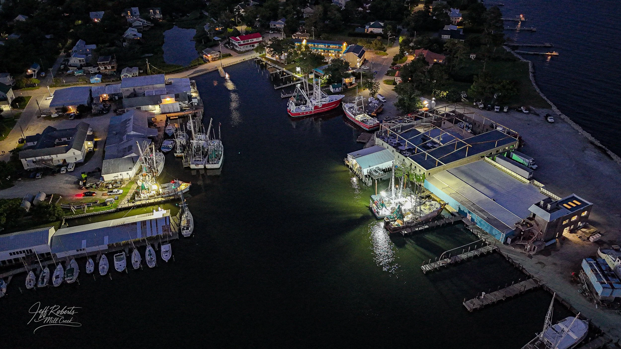 Aerial view of a small harbor at dusk with illuminated boats docked, surrounding buildings, trees, and houses, and a winding shoreline and water neighboring the jetties.