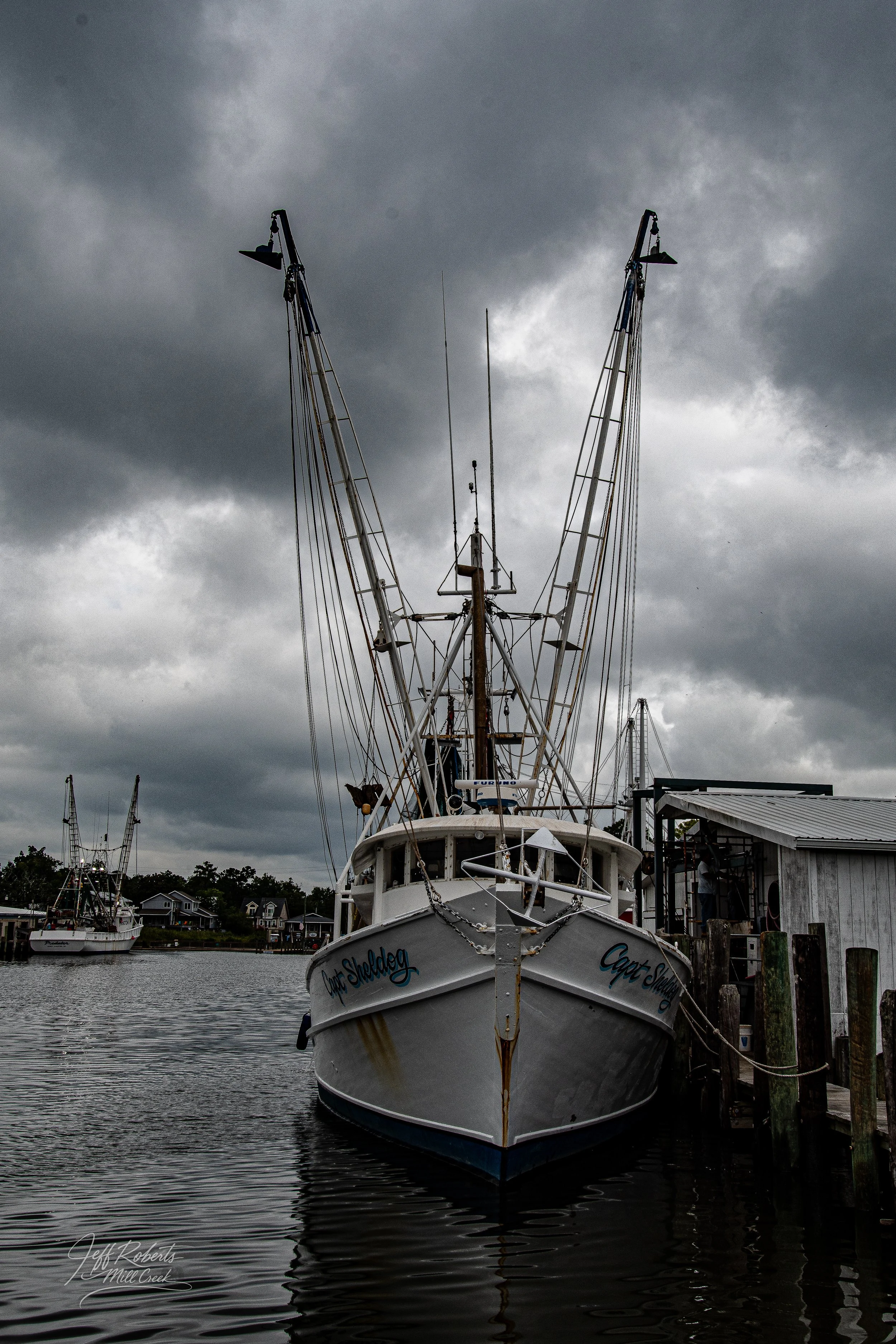 A boat named 'Capt. Sheldog' docked at a pier under a dark, cloudy sky with other boats in the background.