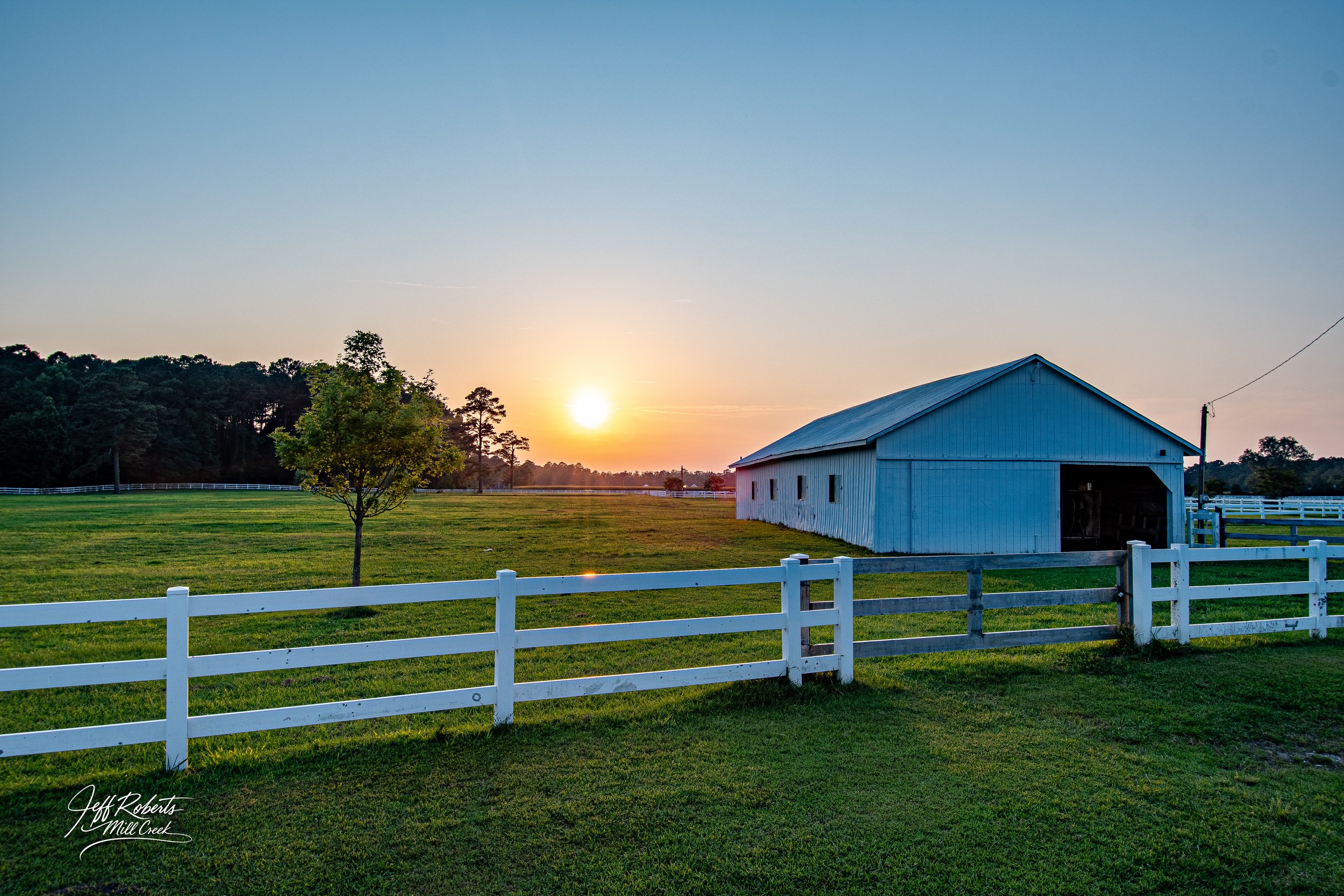 A rural farm scene at sunset with a white barn, a small tree, green grass, white fencing, and a distant tree line under a clear sky.
