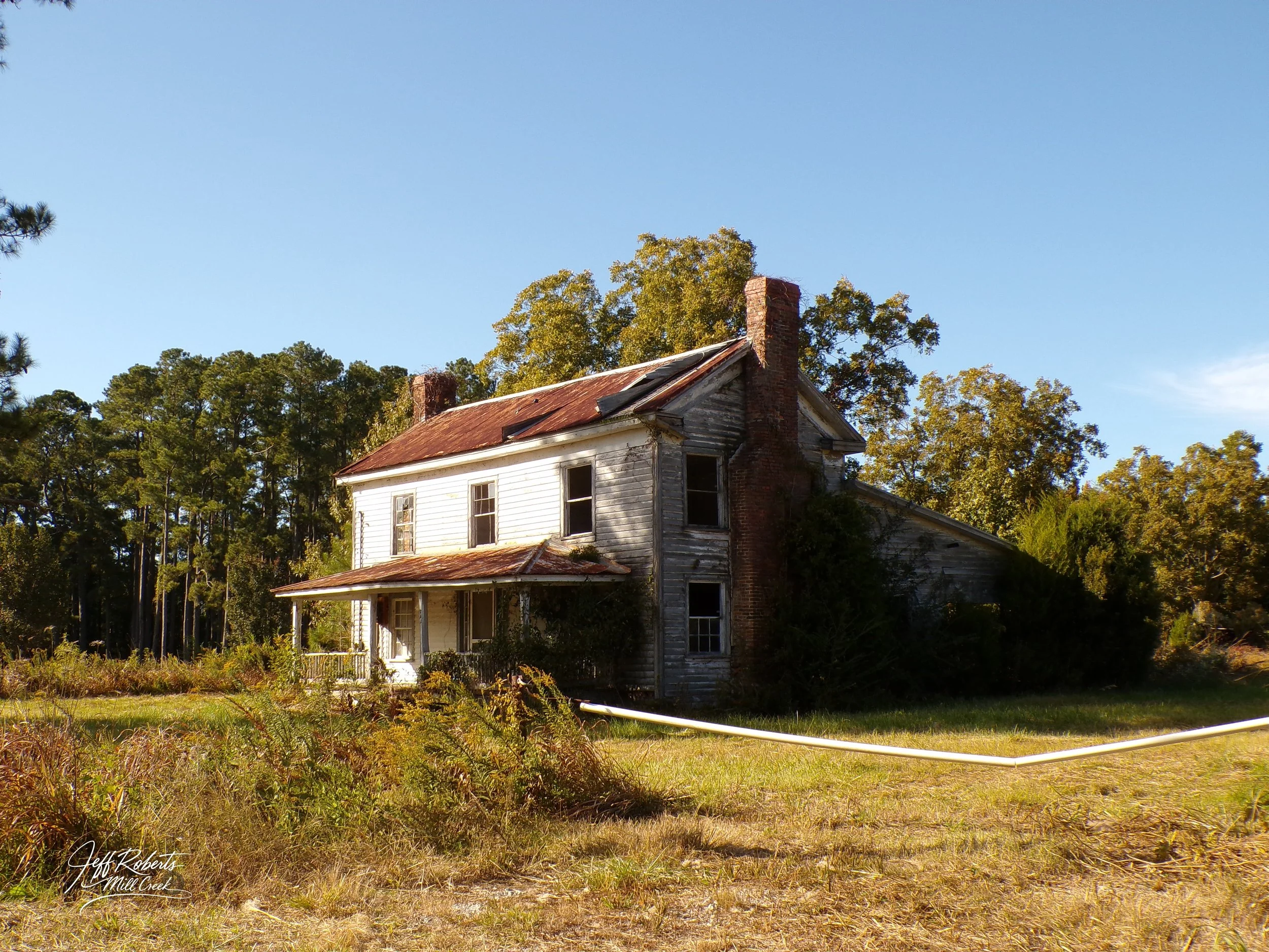 Old, abandoned two-story house with peeling paint, broken windows, rusted roof, and overgrown yard, surrounded by trees under a clear blue sky.
