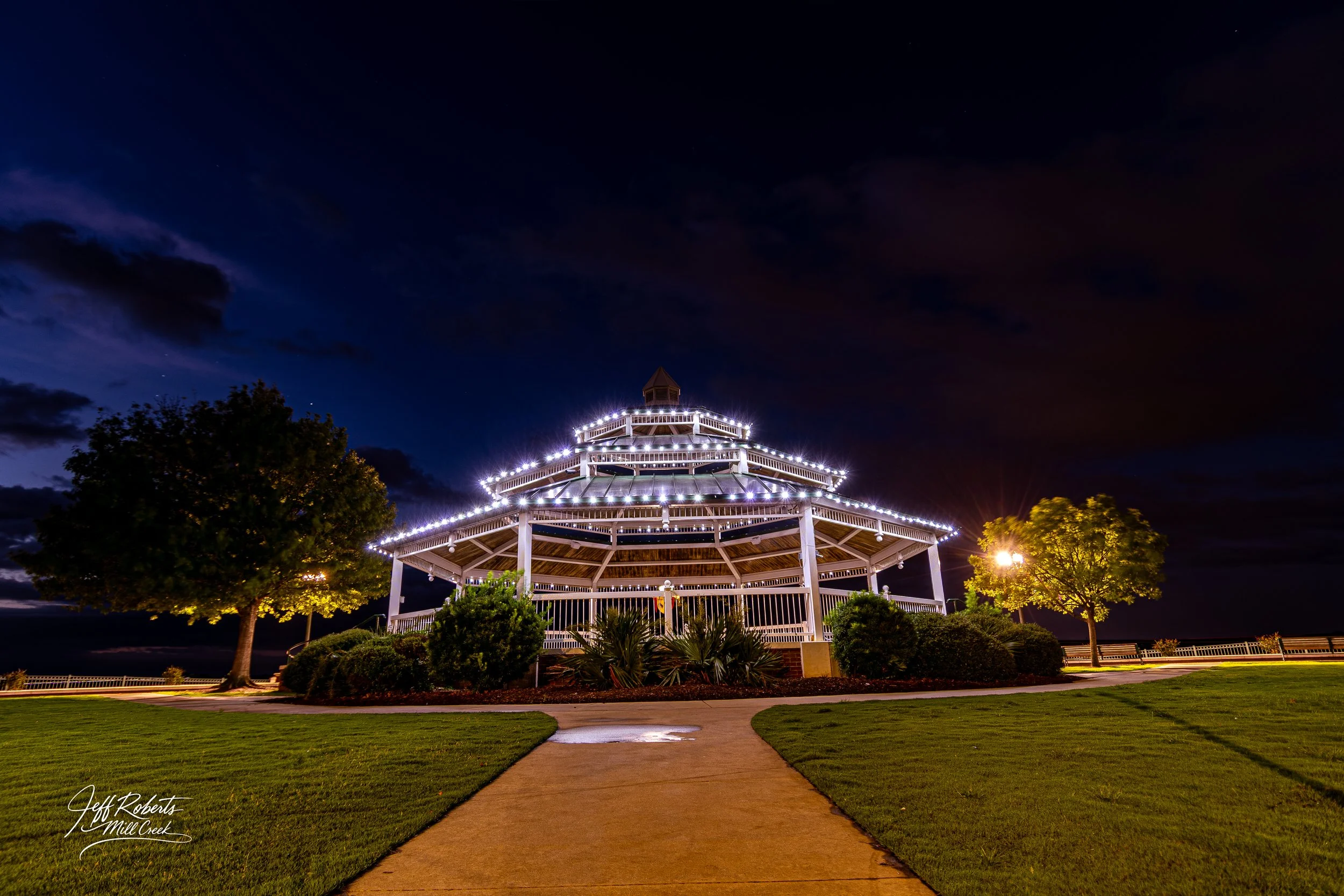 A lit-up, multi-tiered gazebo in a park at night with trees, grass, and a cloudy sky.