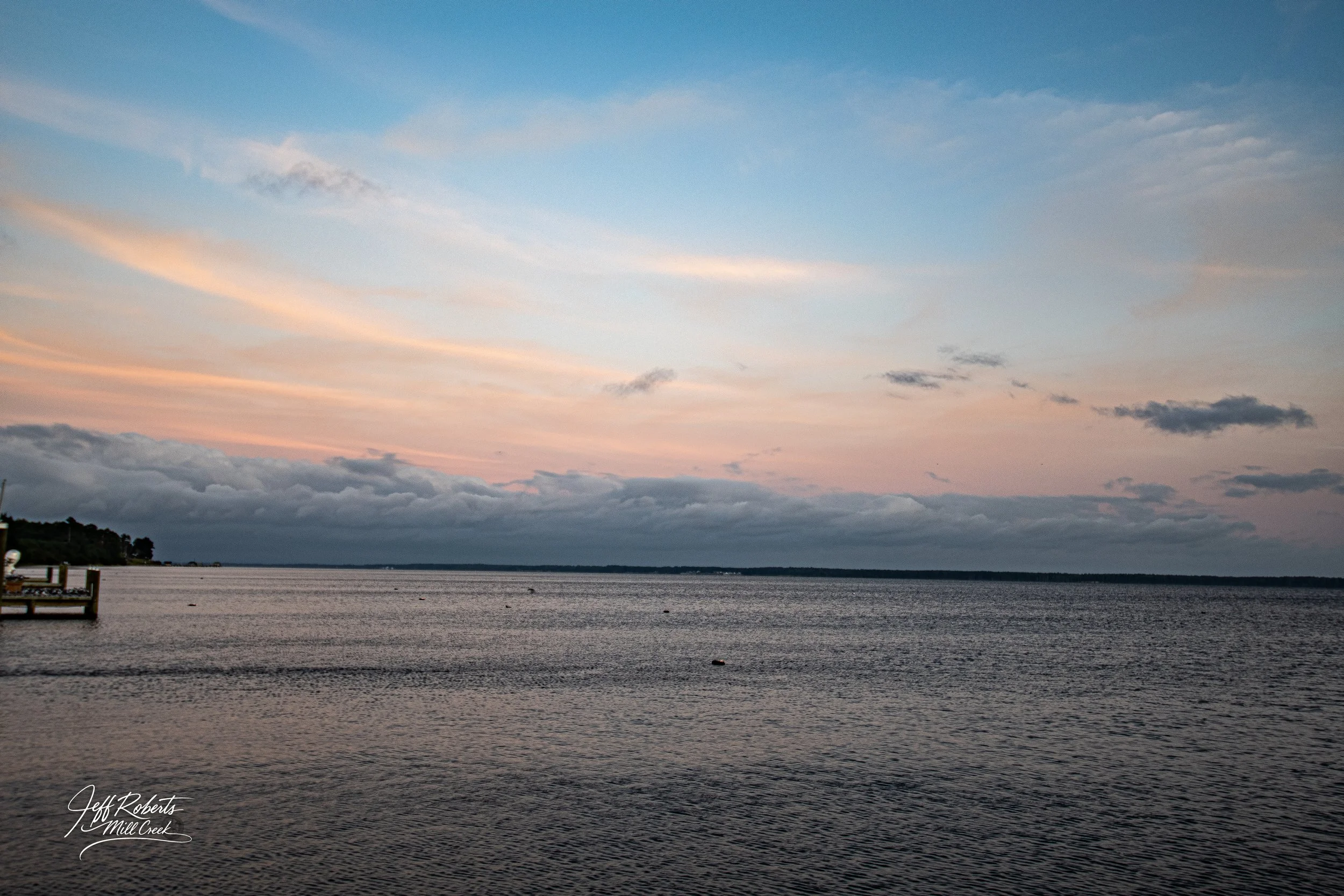 A calm body of water at sunset with a cloudy sky featuring pink and orange hues and a distant shoreline with trees on the horizon, and a small dock extending into the water on the left side.