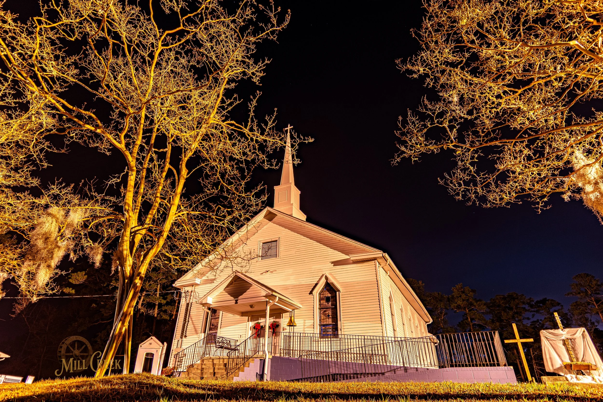 A white church building with a tall steeple, decorated with red wreaths, illuminated at night, surrounded by leafless trees and outdoor crosses.