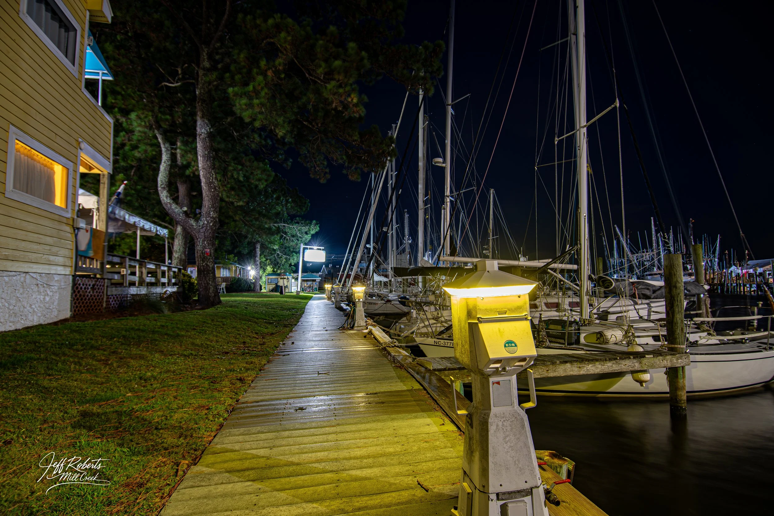 Nighttime scene at a marina with sailboats docked, a wooden sidewalk, trees, a yellow building on the left, and illuminated signage in the distance.