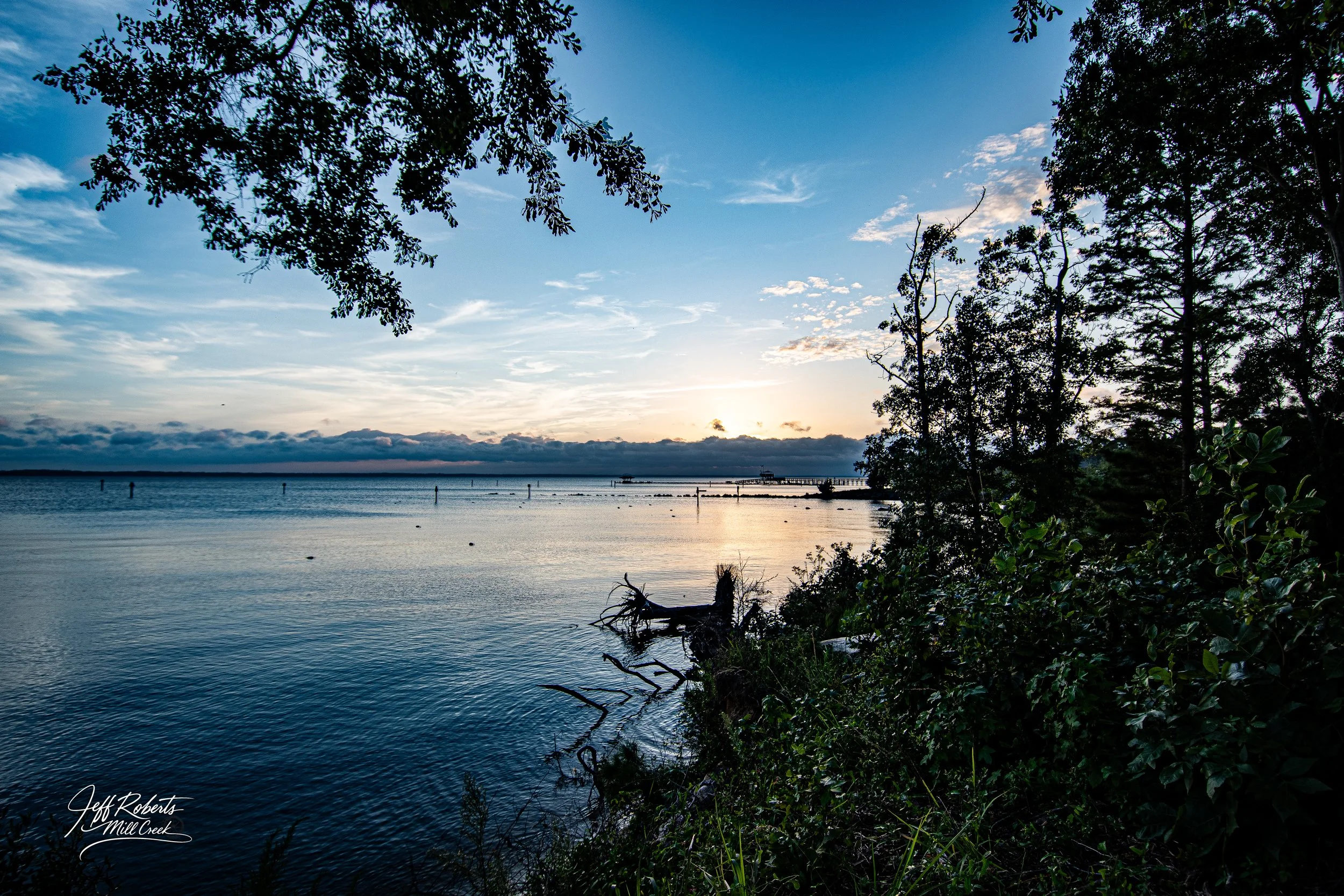 A serene view of Mill Creek at sunset with calm water, silhouetted trees on the right, and a partially cloudy sky with the sun near the horizon.
