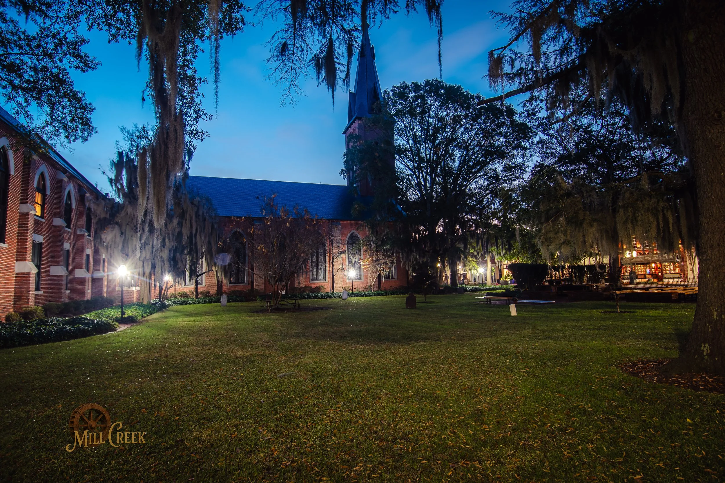 A church with a tall steeple, brick walls, and large arched windows, surrounded by trees with Spanish moss, illuminated by outdoor lights at dusk.