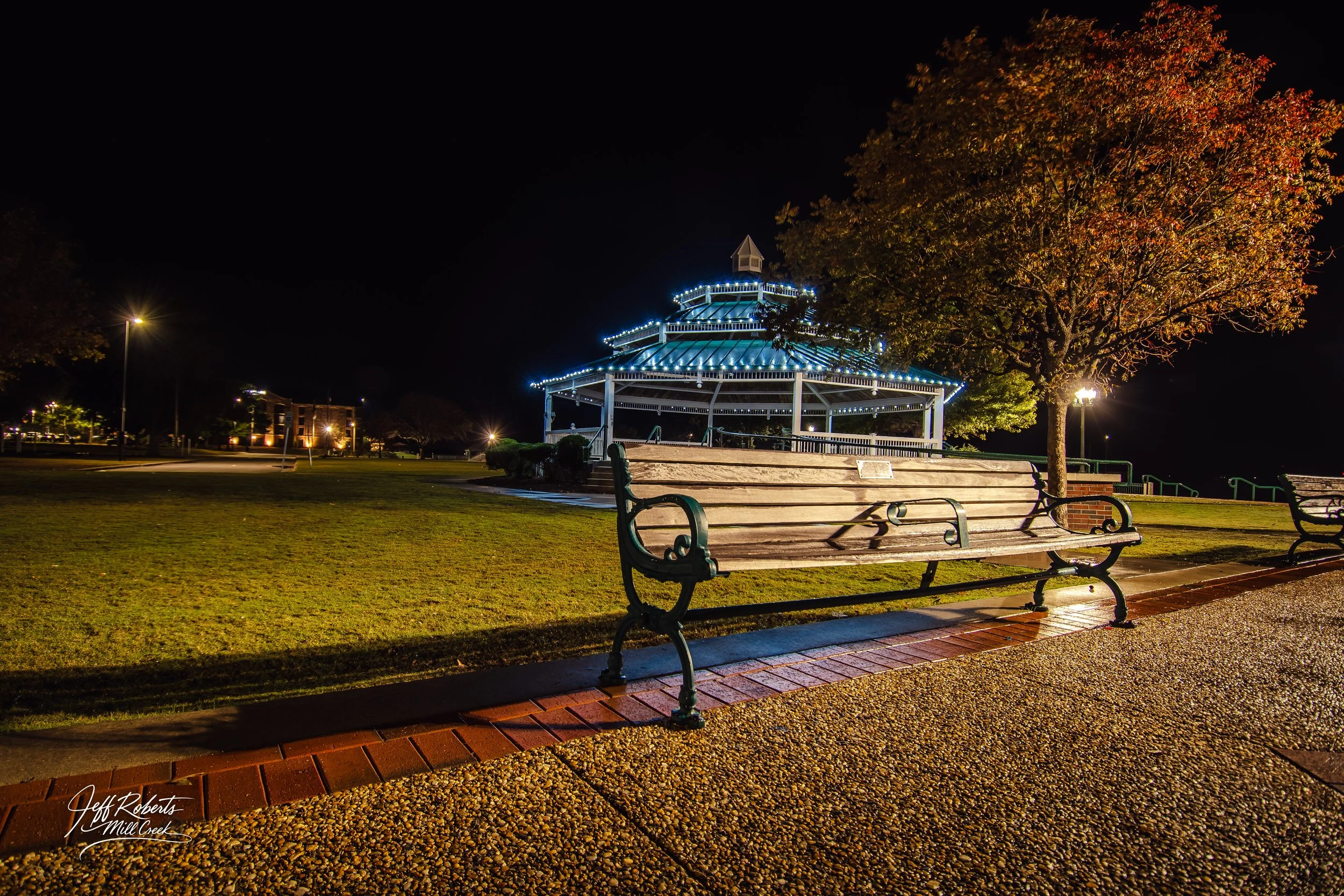 Night view of a park with a lit pavilion, benches, and trees, illuminated by streetlights.