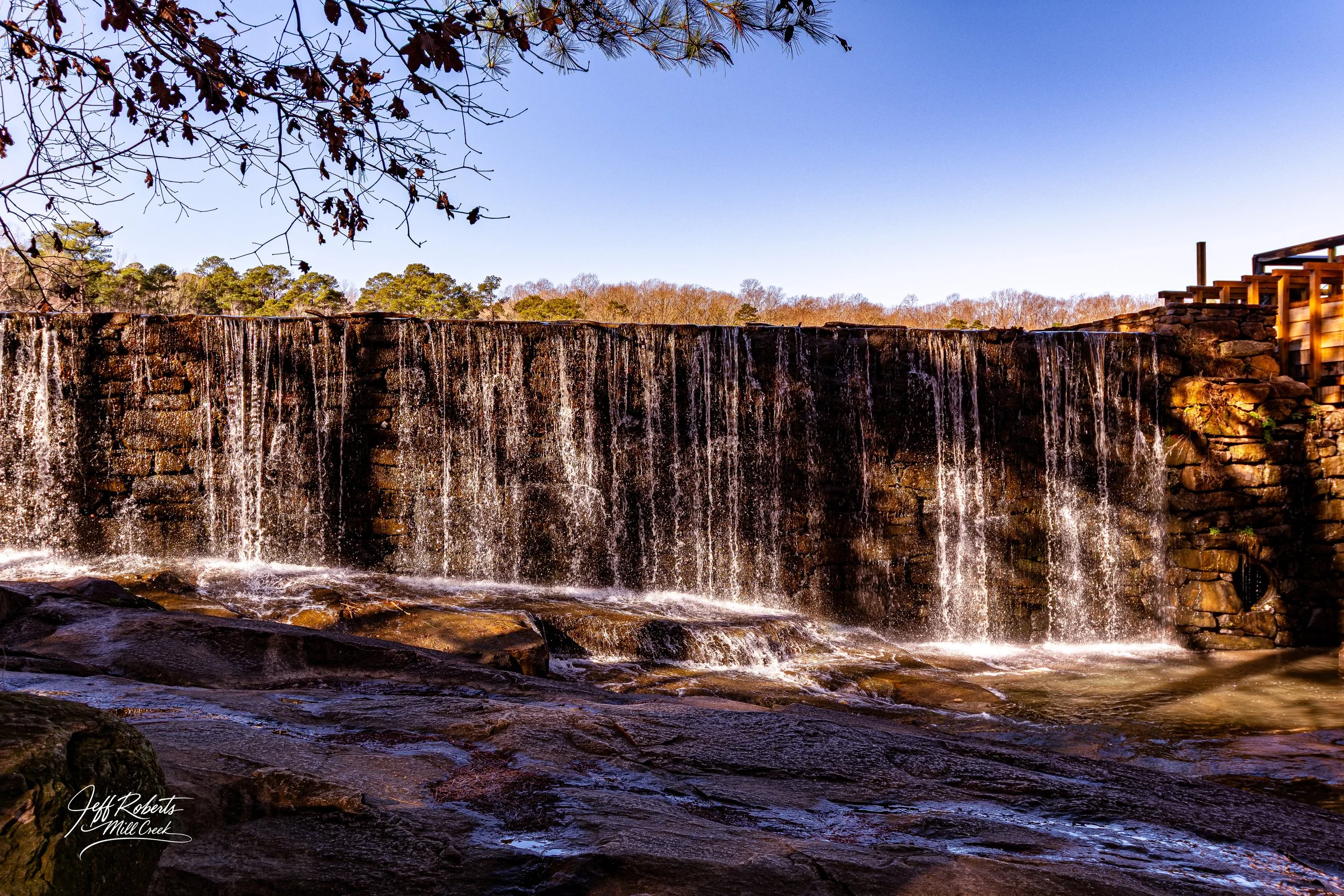 Waterfall over a stone dam with clear water at the bottom, trees and blue sky in the background, and overhanging branches at the top.