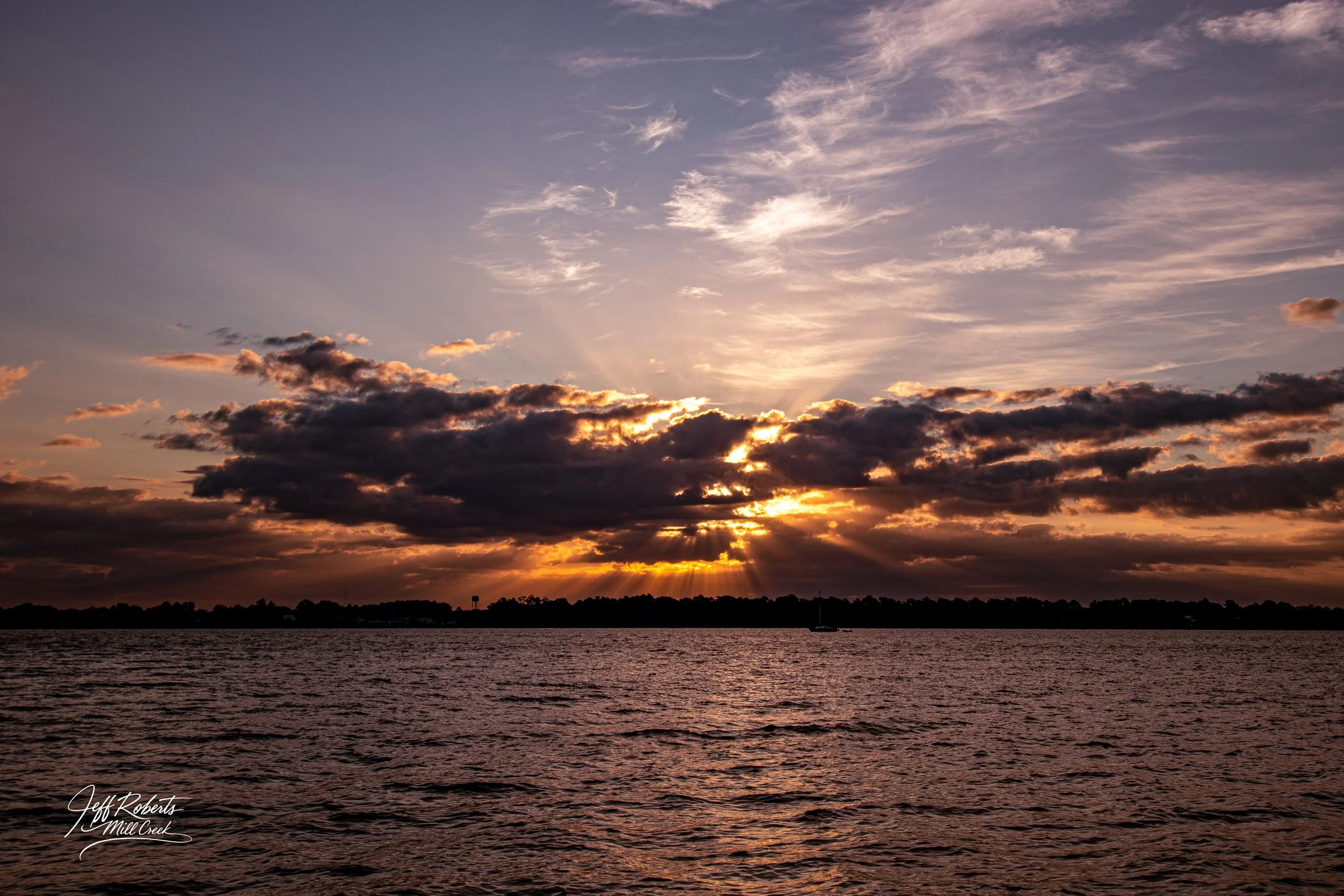 A sunset over a body of water with clouds partially obscuring the sun and rays of light shining through, silhouetted trees and landscape on the horizon.