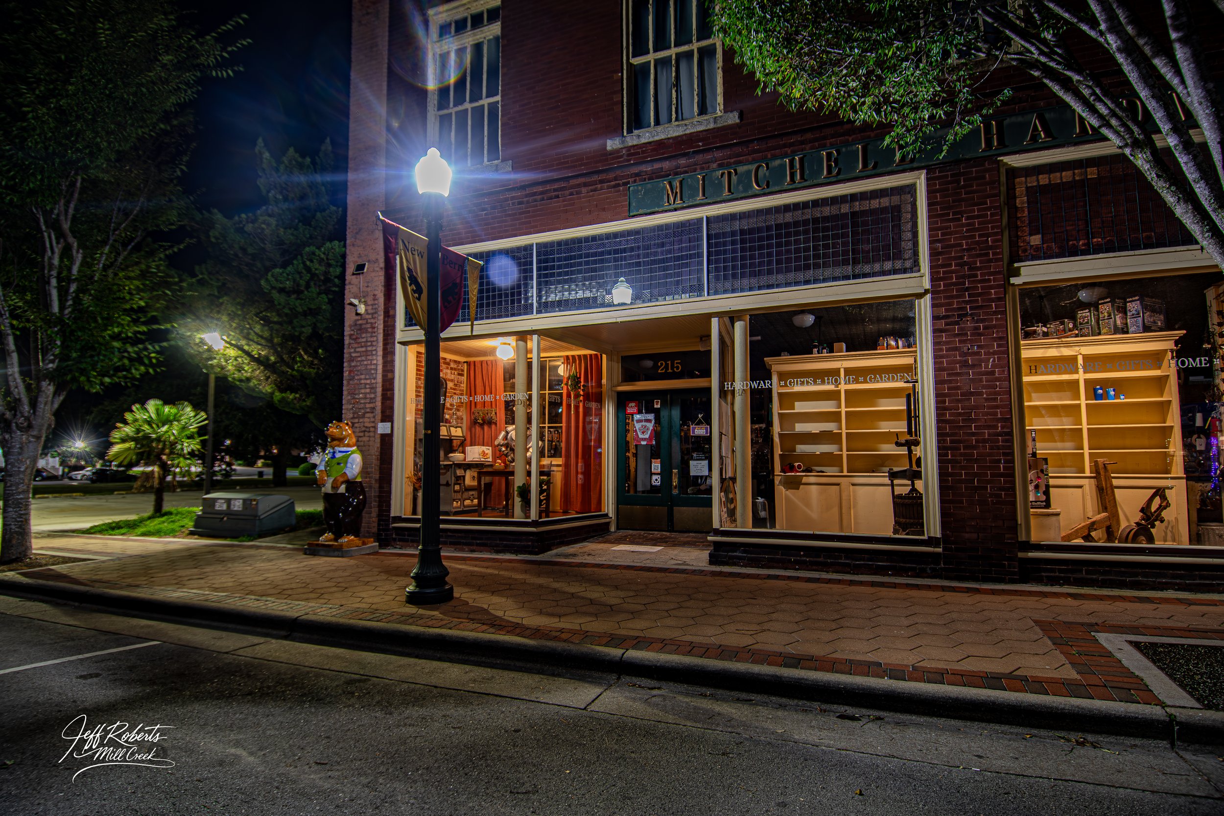 Night scene of a brick storefront with yellow shelving inside and a sign that reads "MITCHEL". A street lamp and a bear statue are visible outside, along with trees and parked cars in the background.