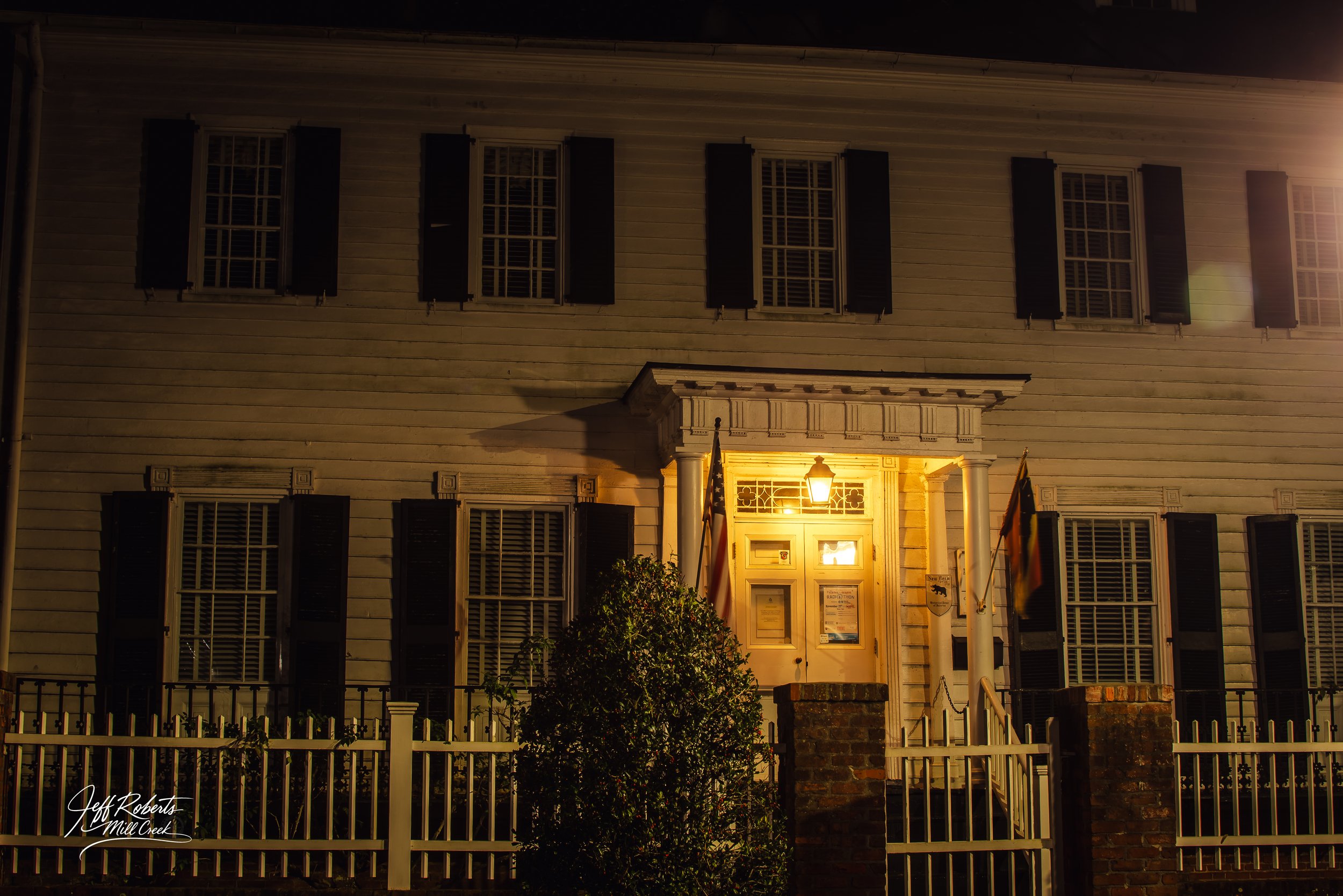 A white wooden house with black shutters, illuminated by a yellow porch light at night. The house has multiple windows and a small front porch with a brick and white picket fence.