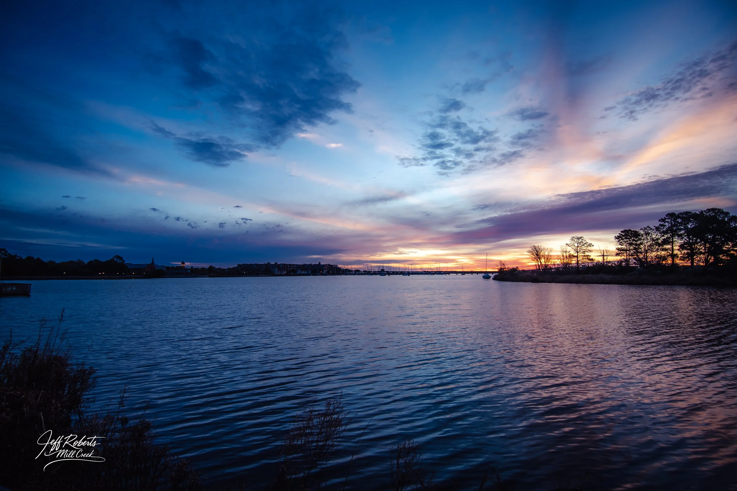 A peaceful river scene at sunset with colorful clouds in the sky, trees along the shoreline, and a few boats anchored on the water.