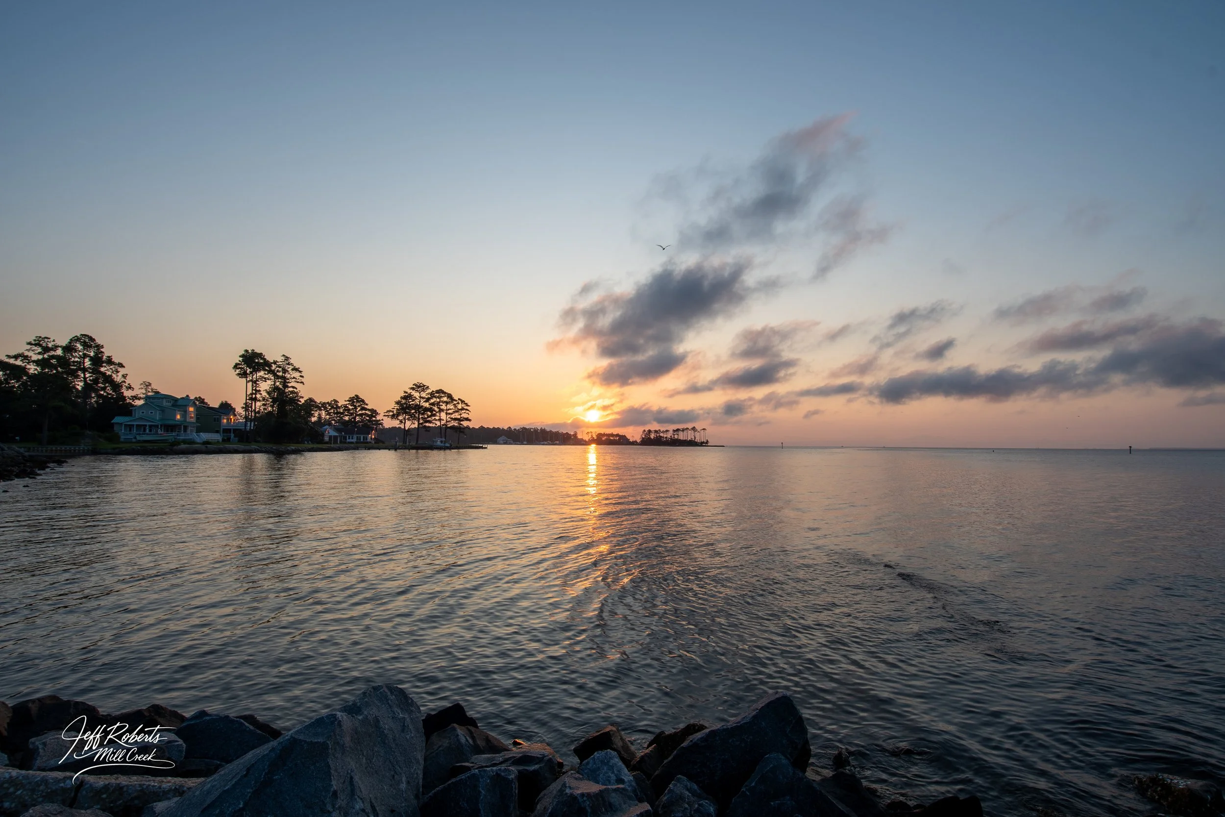 Sunset over a calm body of water with a shoreline of rocks in the foreground, trees and houses on the left, scattered clouds in the sky, and a bird flying overhead.