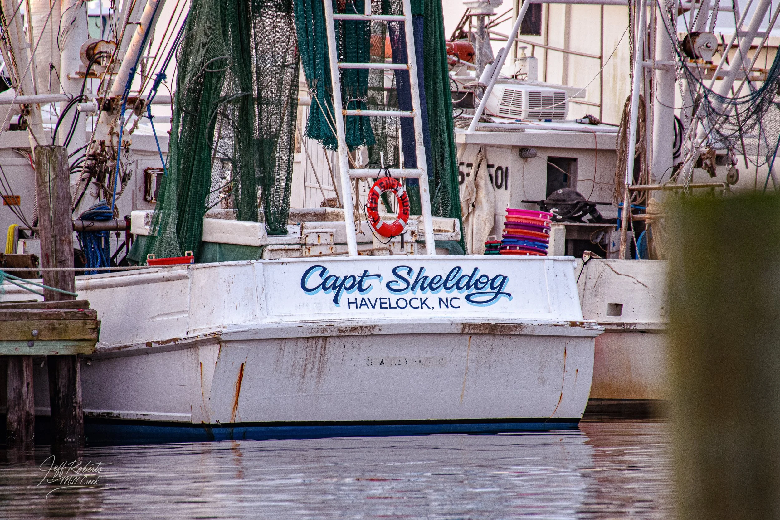 A white fishing boat named 'Capt Shel Dog' docked at a marina in Havelock, North Carolina, with fishing nets and equipment on board.