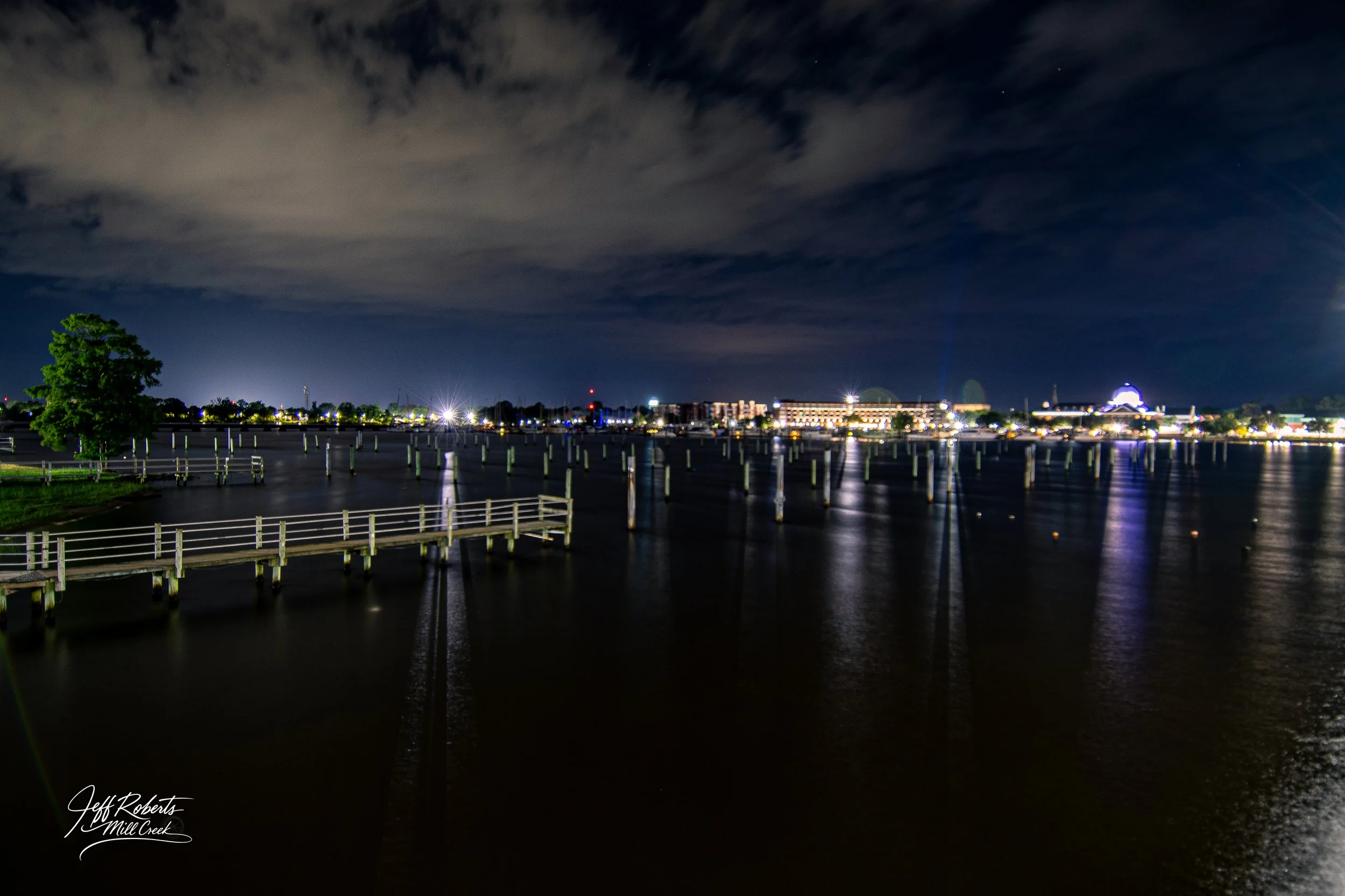 Night view of a city waterfront with illuminated buildings and docks, cloudy sky, and reflections on dark water.