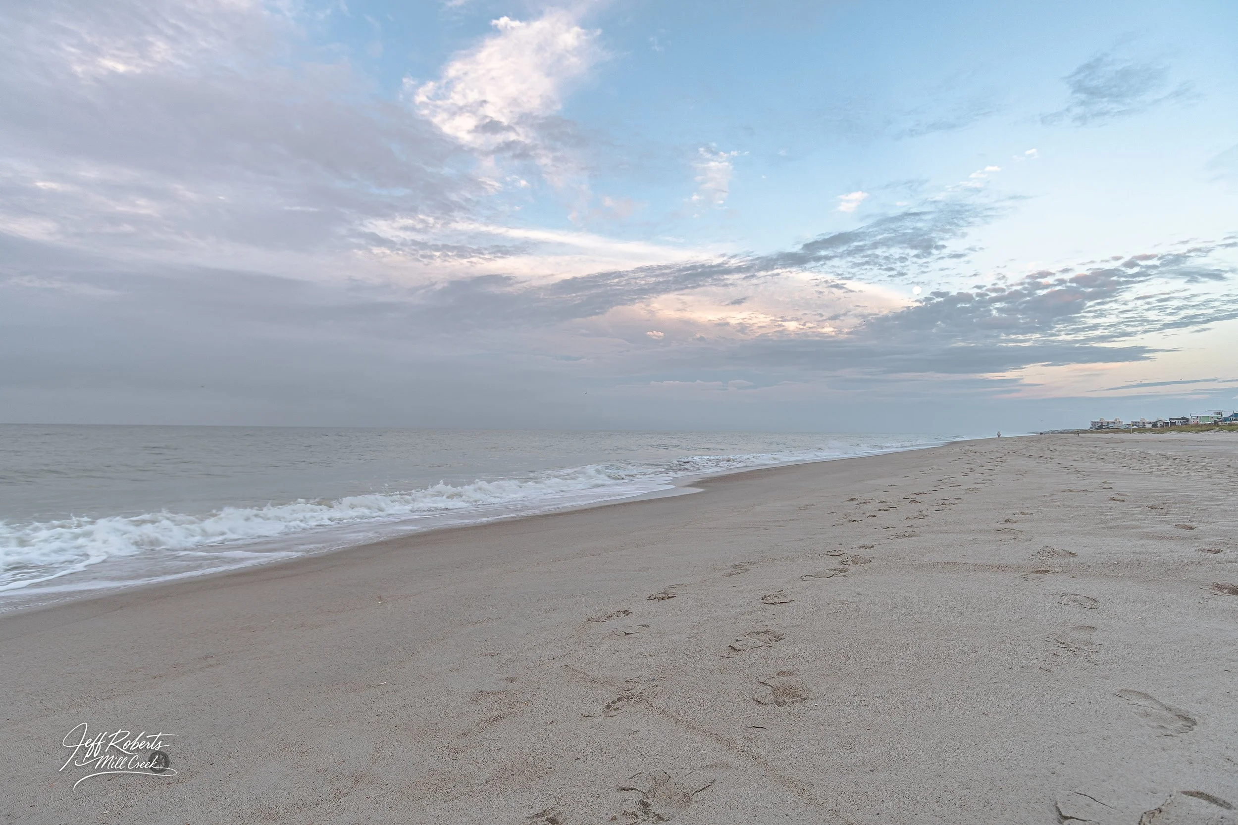 Beach with footprints in the sand, ocean waves, cloudy sky during sunset or sunrise, and distant buildings on the horizon.