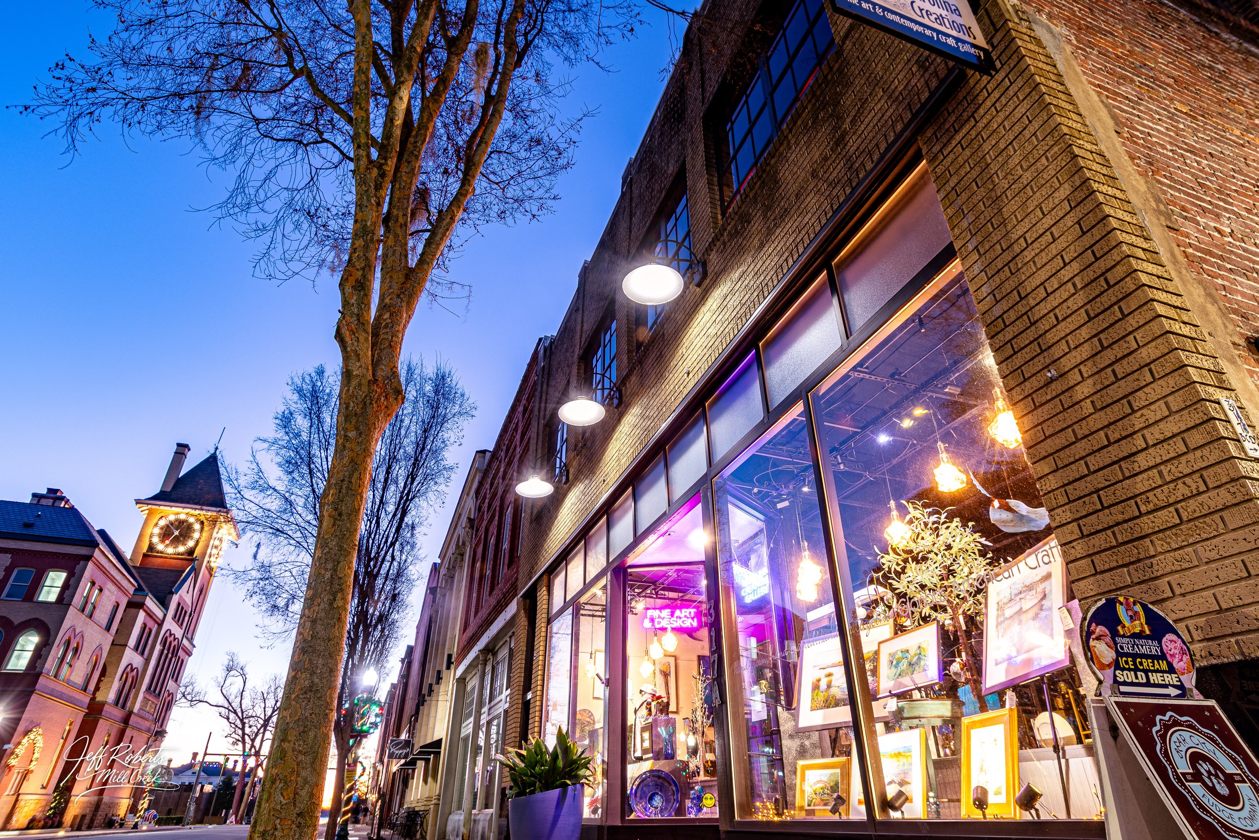 A street scene at dusk with a brick building featuring large glass windows displaying artwork and illuminated by warm lighting inside. A leafless tree in the foreground, street lamps, and historic buildings with clock towers are visible.