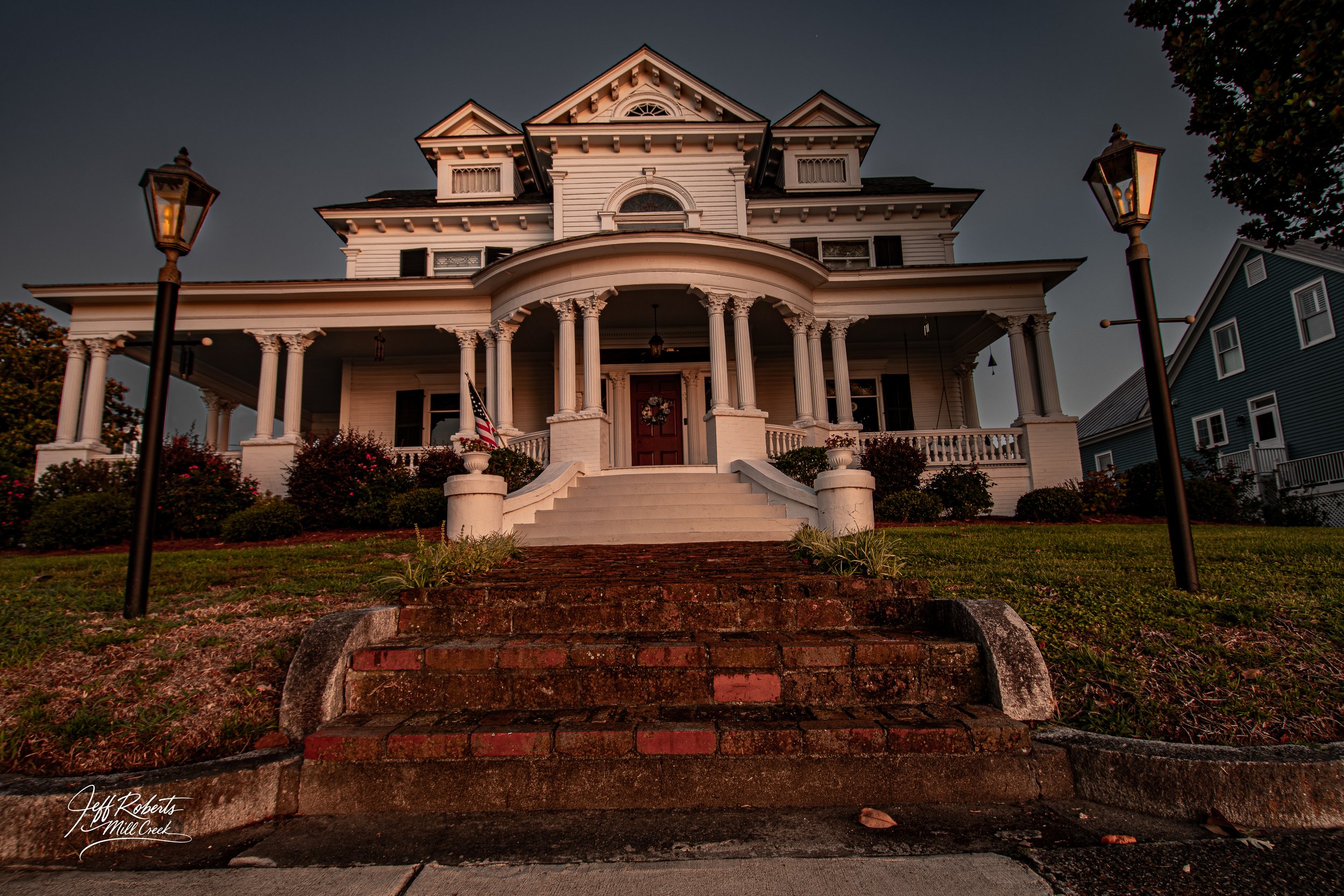 Historic white house with tall columns, steps leading to a red front door with a wreath, surrounded by landscaping and lamp posts.