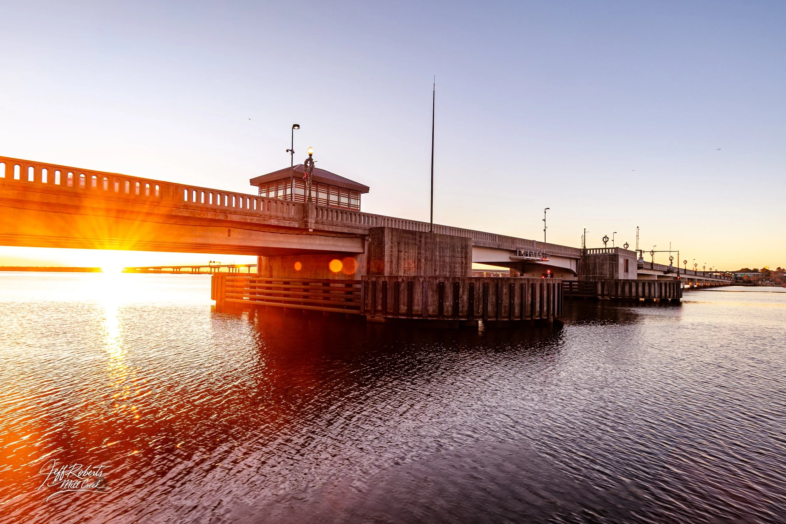 Sunset over a bridge spanning a body of water with reflections and a small building on the bridge