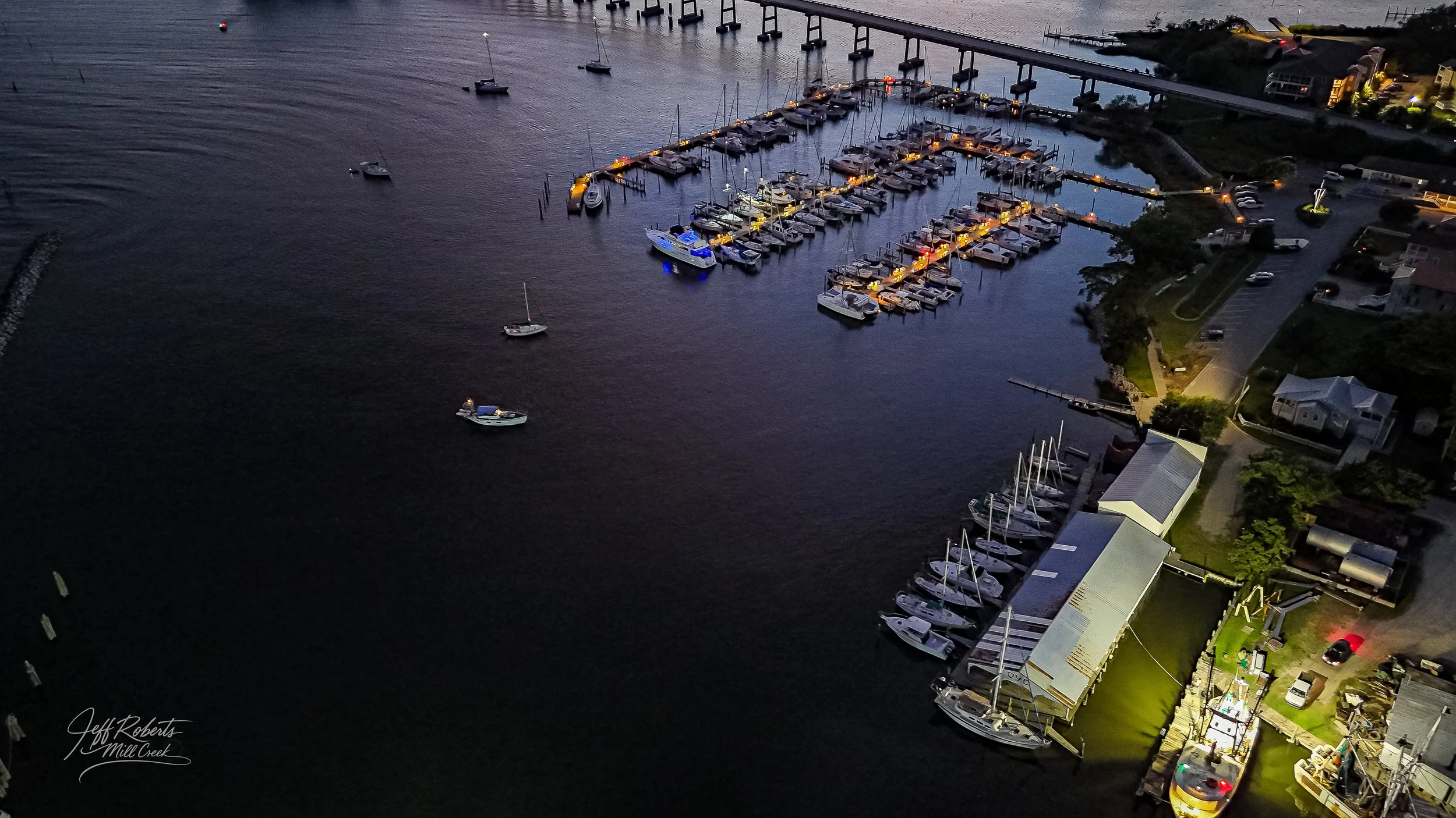 Aerial view of a marina at dusk with boats docked along piers, a parking lot, and buildings nearby, illuminated by streetlights and boat lights.