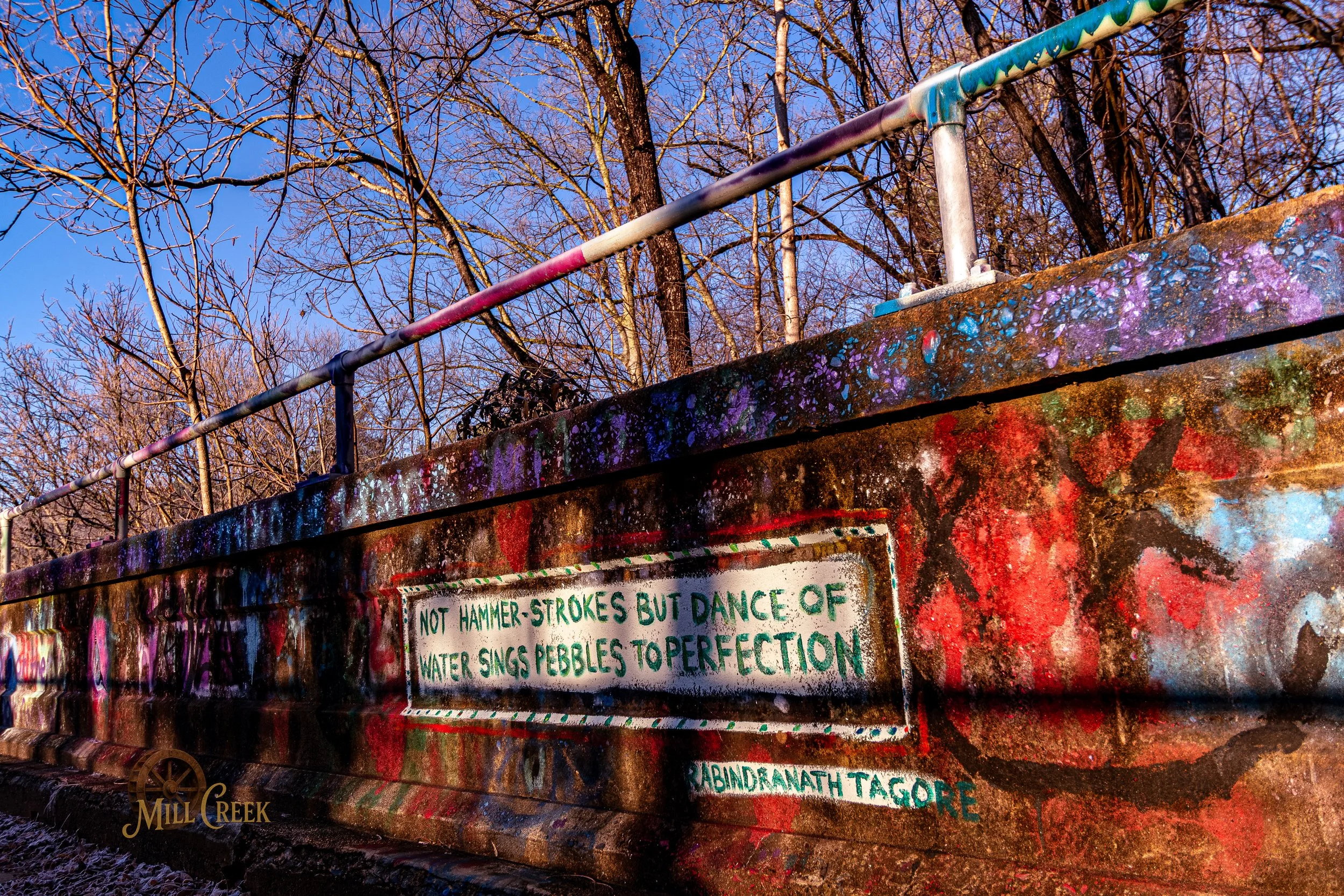 Colorful graffiti-covered concrete structure with a painted quote and trees in the background, under a blue sky.