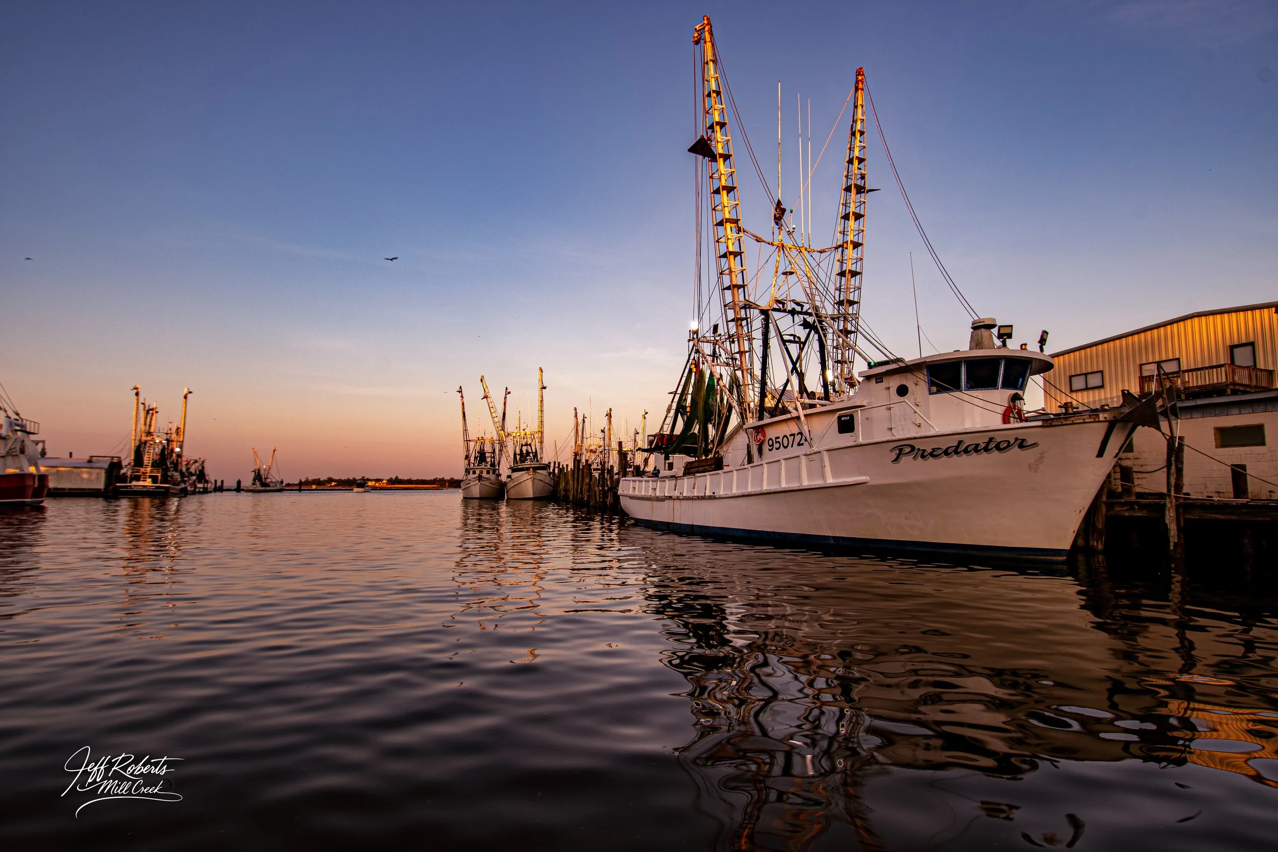 Boats docked at a harbor during sunset, with calm water reflecting the boats and a clear sky.