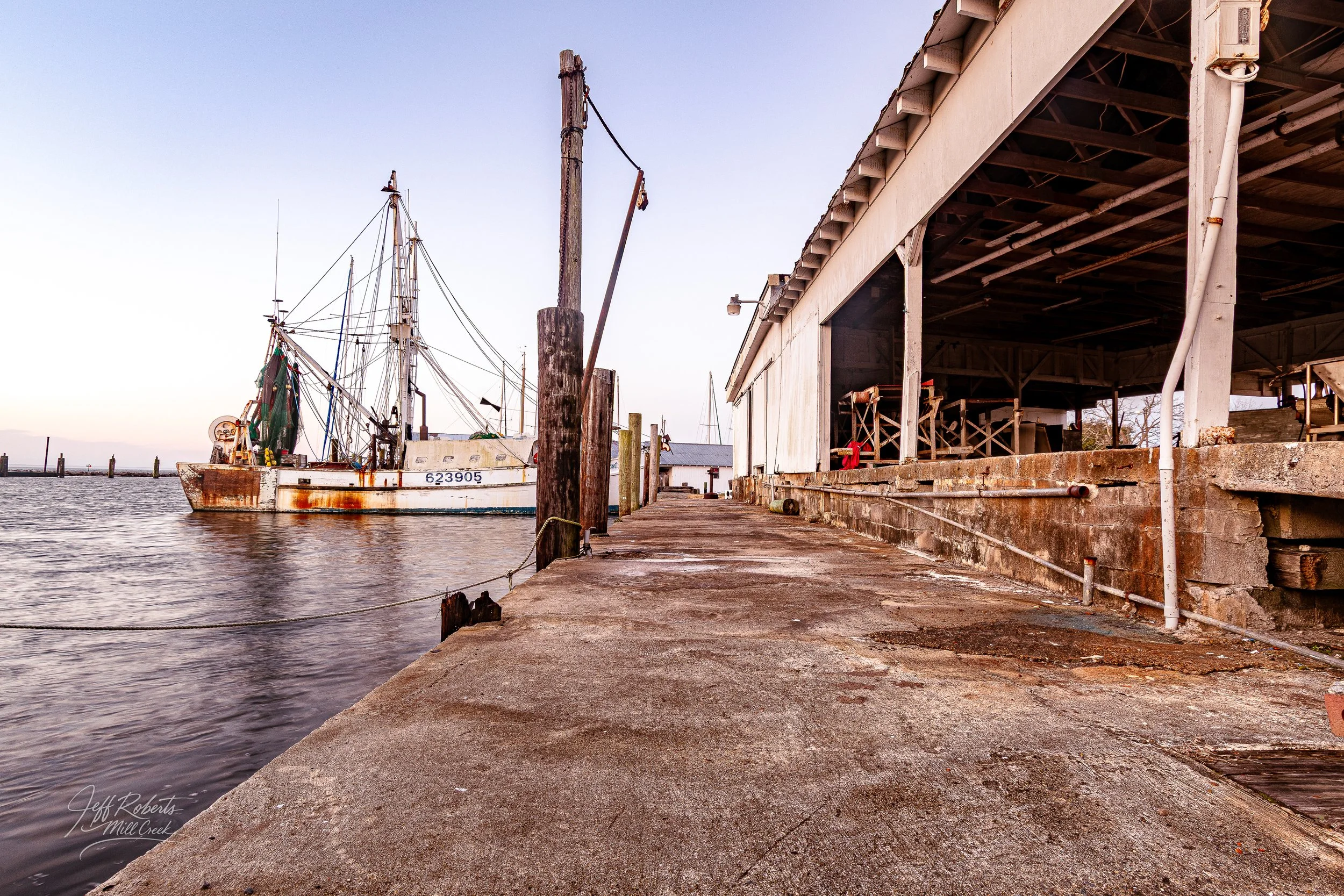 Dockside view with a rusty boat moored on the water, alongside a weathered, open-sided shed on a concrete walkway at a marina.