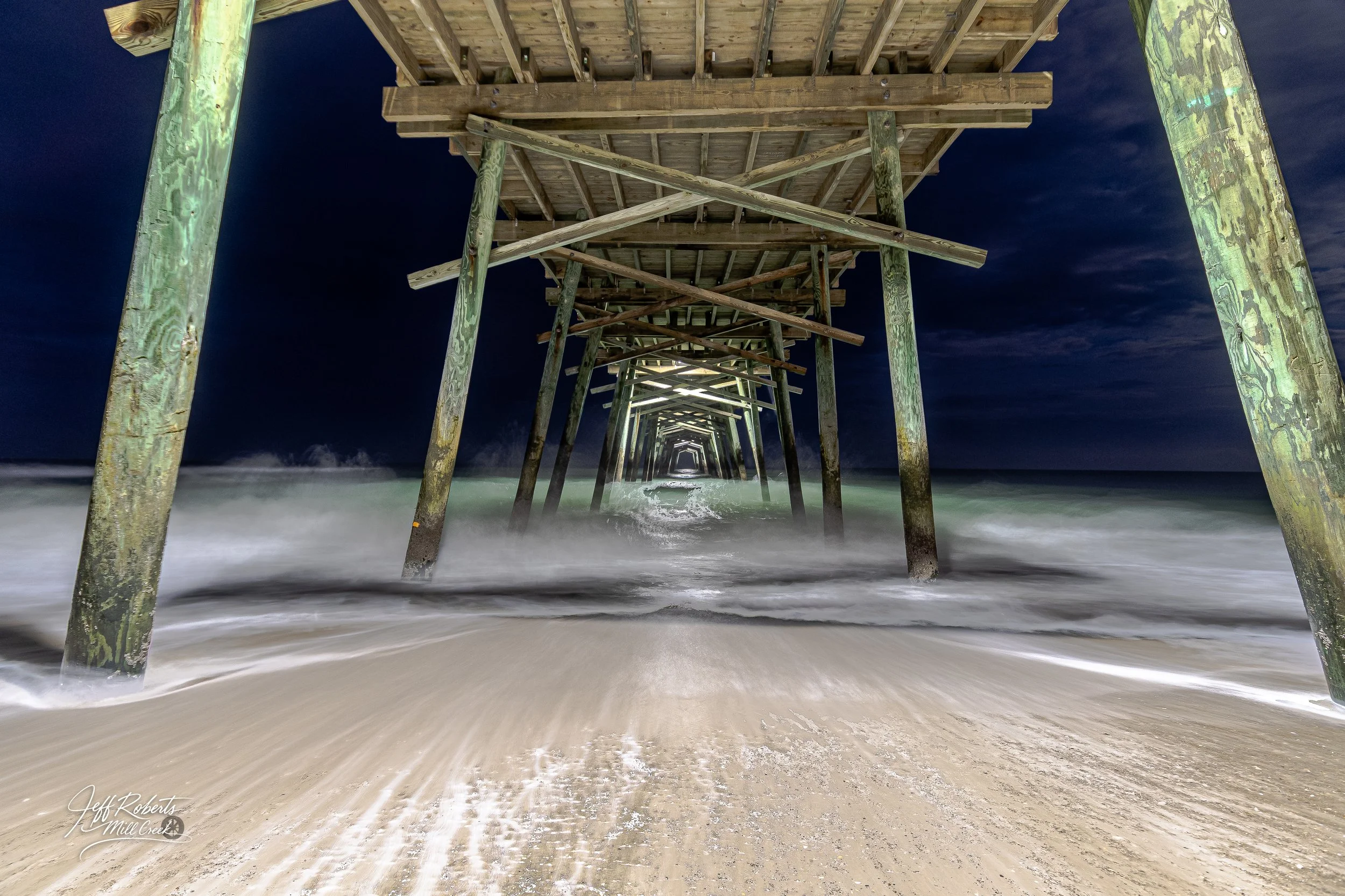 Night view of a wooden pier extending over the ocean, with waves washing onto the sandy beach beneath it.