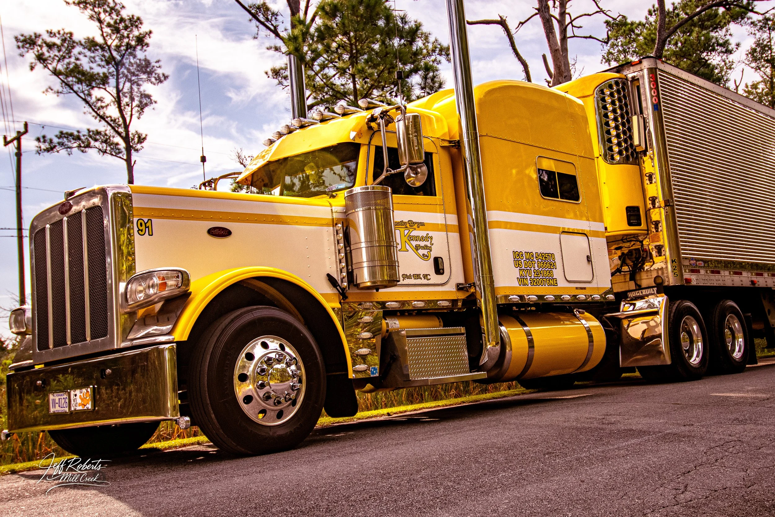 A large yellow semi-truck with chrome accents parked on a paved road next to grassy area with trees in the background.