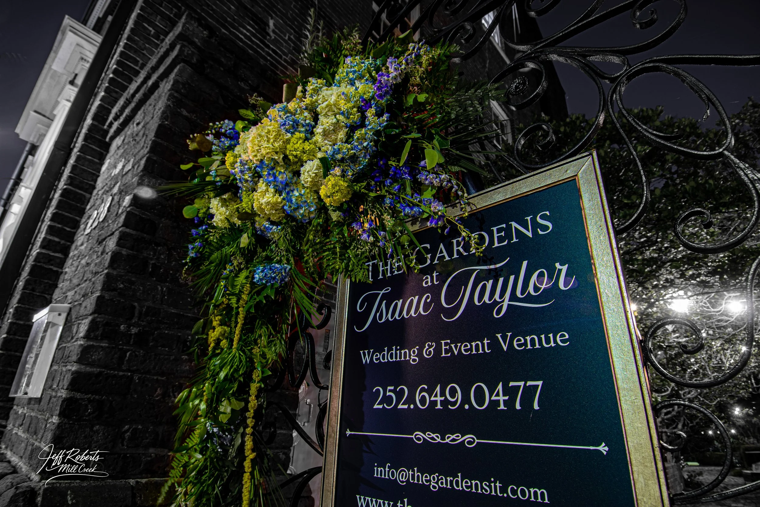 Outdoor event venue sign for The Gardens at Isaac Taylor decorated with a floral arrangement of yellow, blue, and purple flowers, attached to a black wrought iron gate against a brick wall at night.