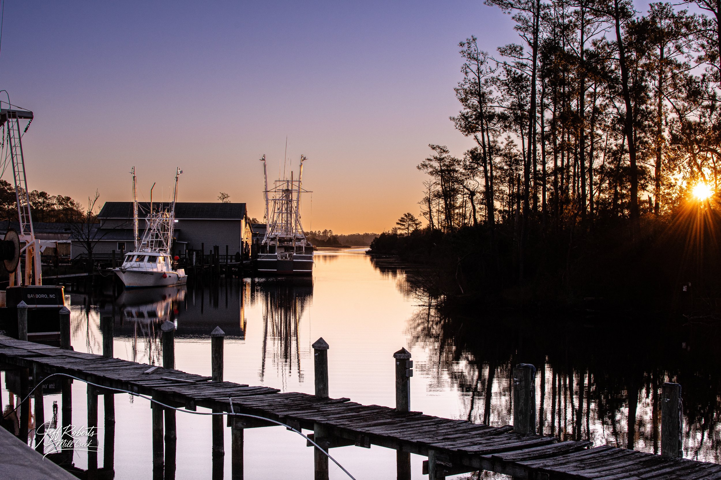 A peaceful waterway with boats docked at a pier, trees lining the shore, and the sun setting in the background.