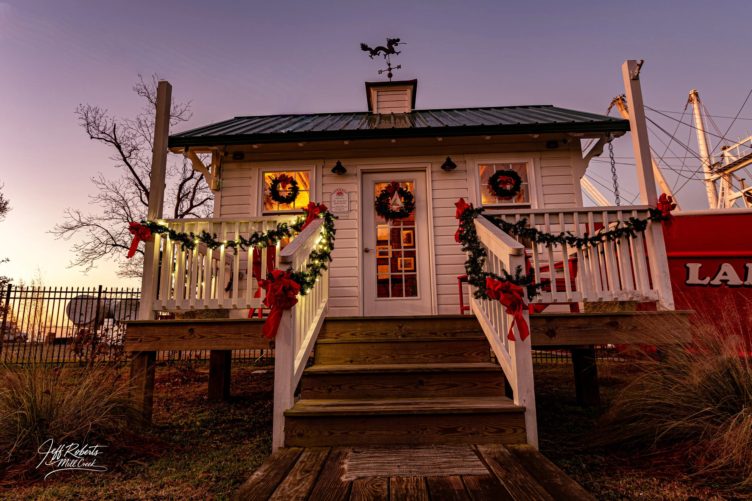 Small white wood house decorated with Christmas wreaths, garlands, and bows, with a staircase leading to the door, at sunset.