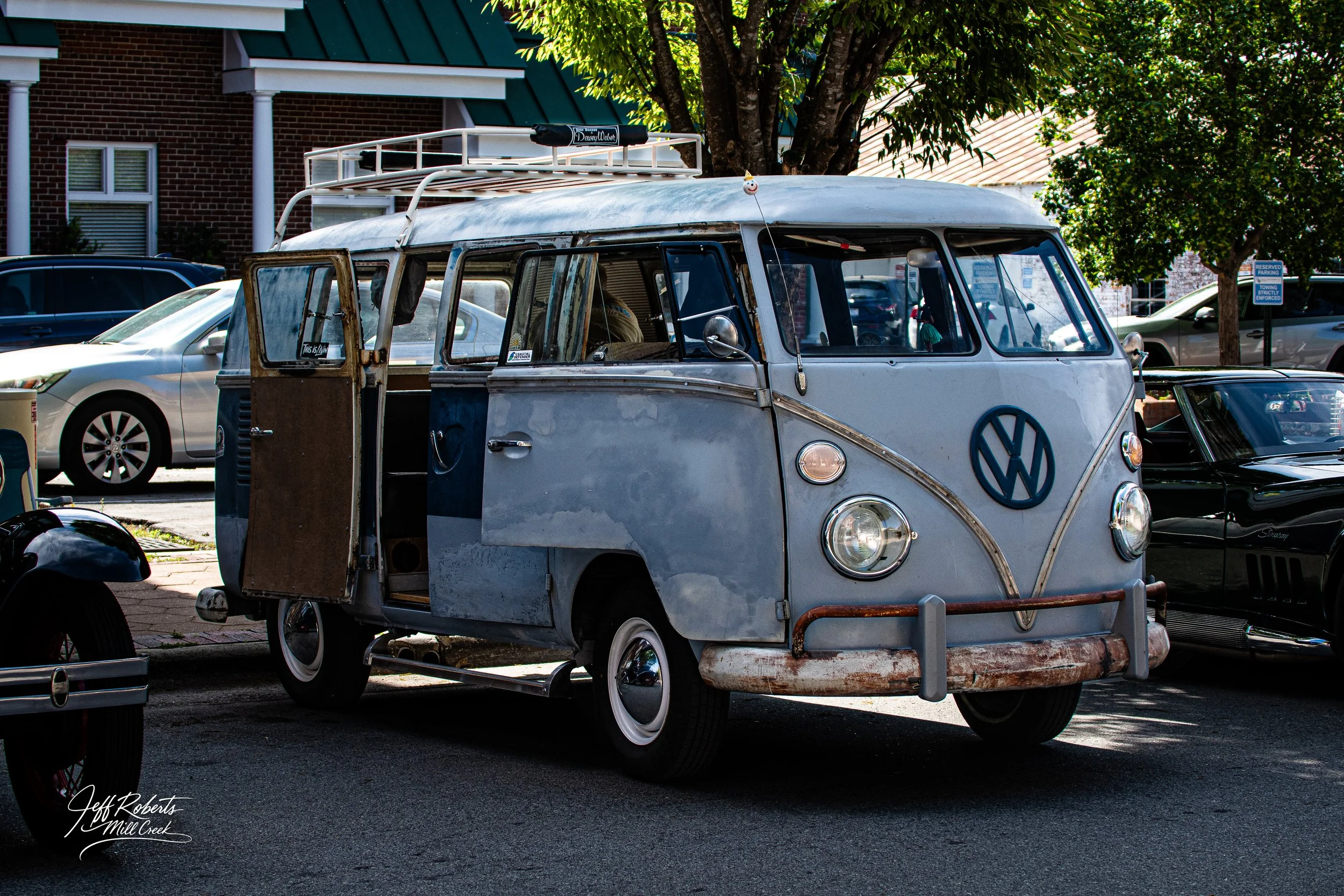 An old Volkswagen Type 2 van with a rusted bumper and weathered paint parked in a parking lot. The van has an open sliding door, a roof rack, and is situated near trees and other modern vehicles.